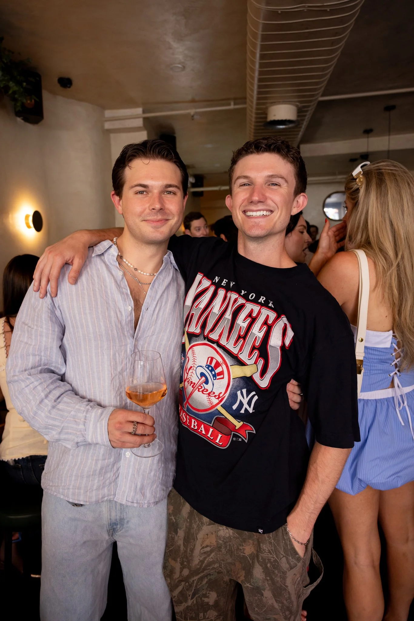 Two young men smiling and posing together at a social gathering; one holding a glass of rosé wine, with people socializing in the background.