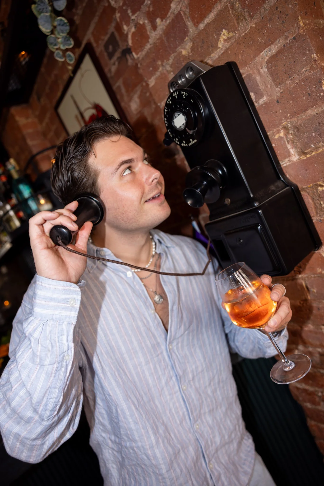 Man in striped shirt talking on vintage black rotary phone mounted on brick wall, holding glass of rosé wine, smiling and looking up.