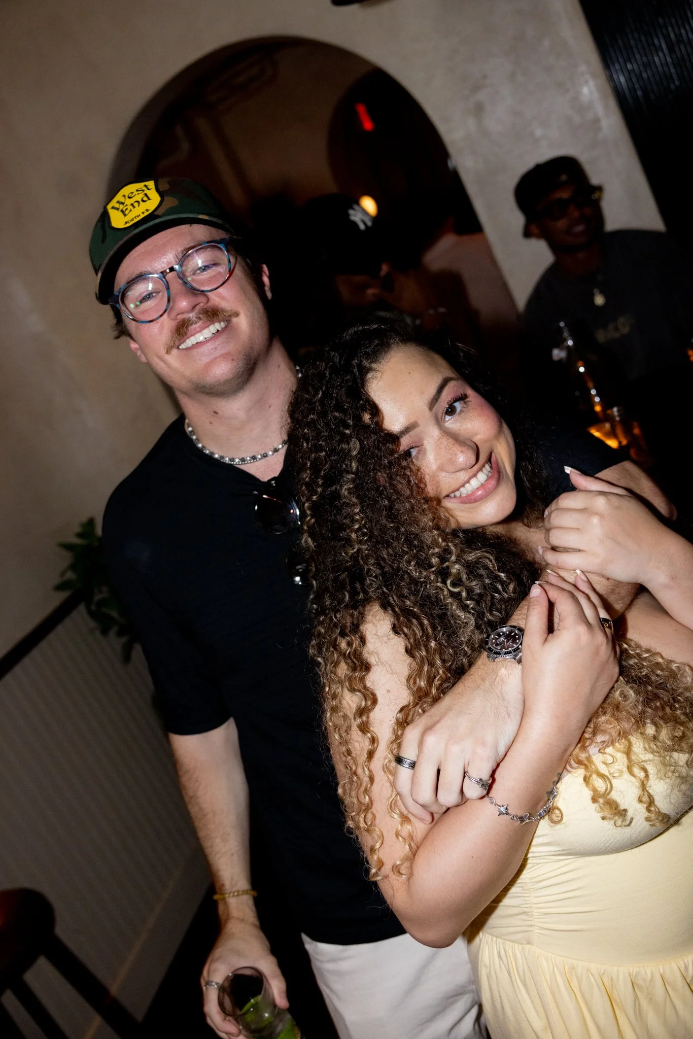 A man and woman smiling at a party, with the man wearing a camouflage cap and the woman with curly hair, hugging and holding hands. The background shows other people and a bar area.