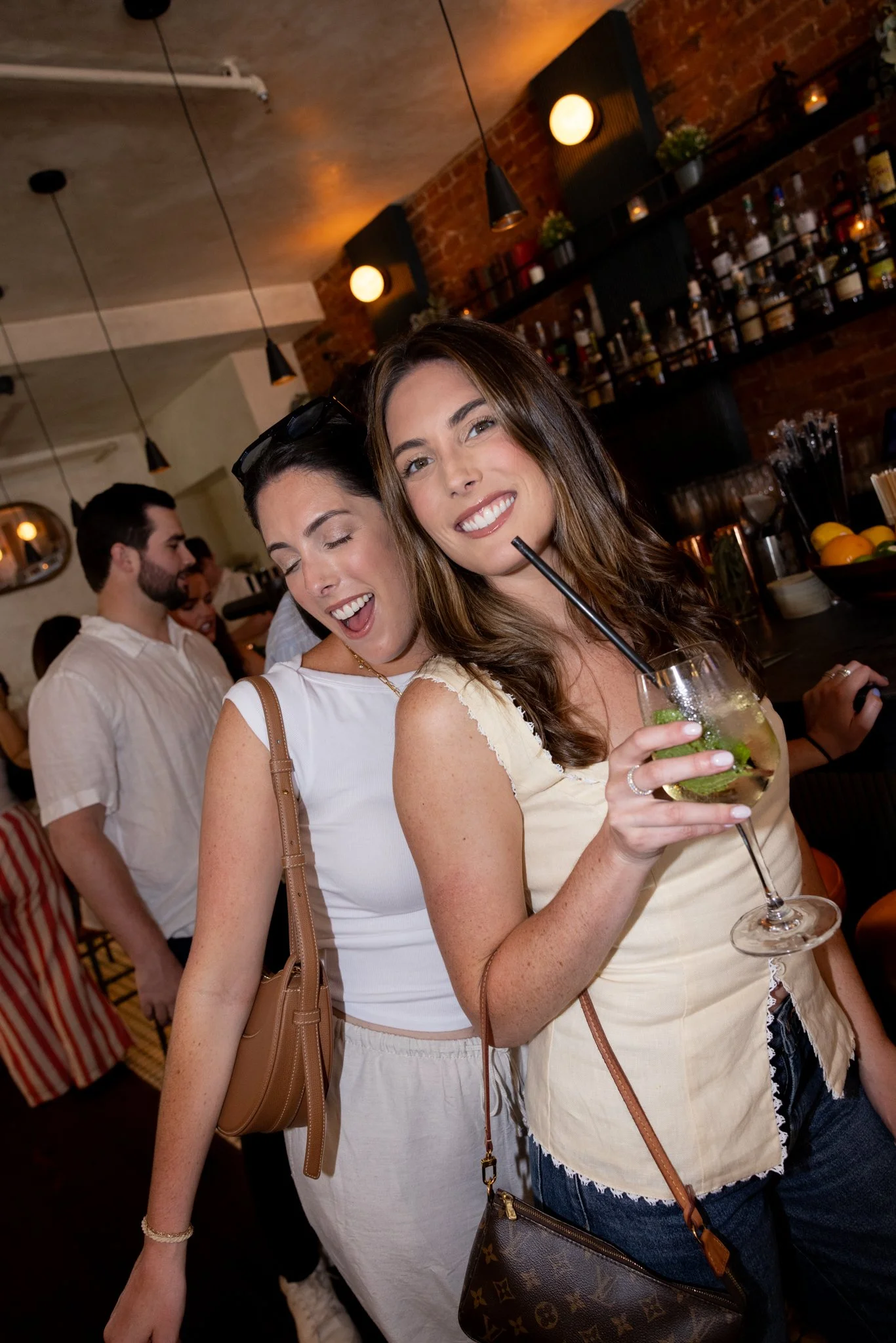 Two women enjoying drinks at a bar, smiling and posing for the camera while others are in the background.