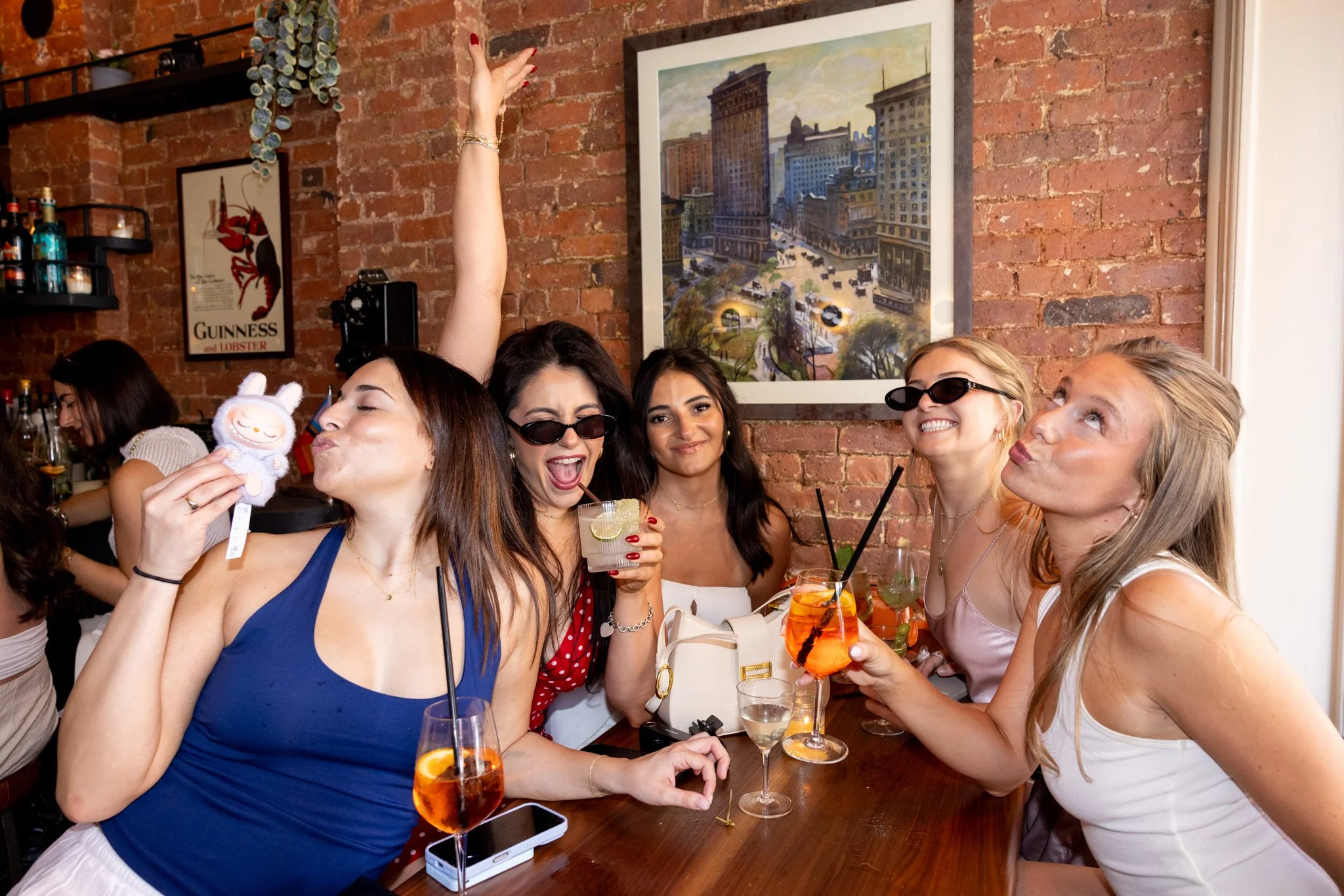 Group of six women enjoying drinks and taking selfies at a party in a bar with a brick wall background.