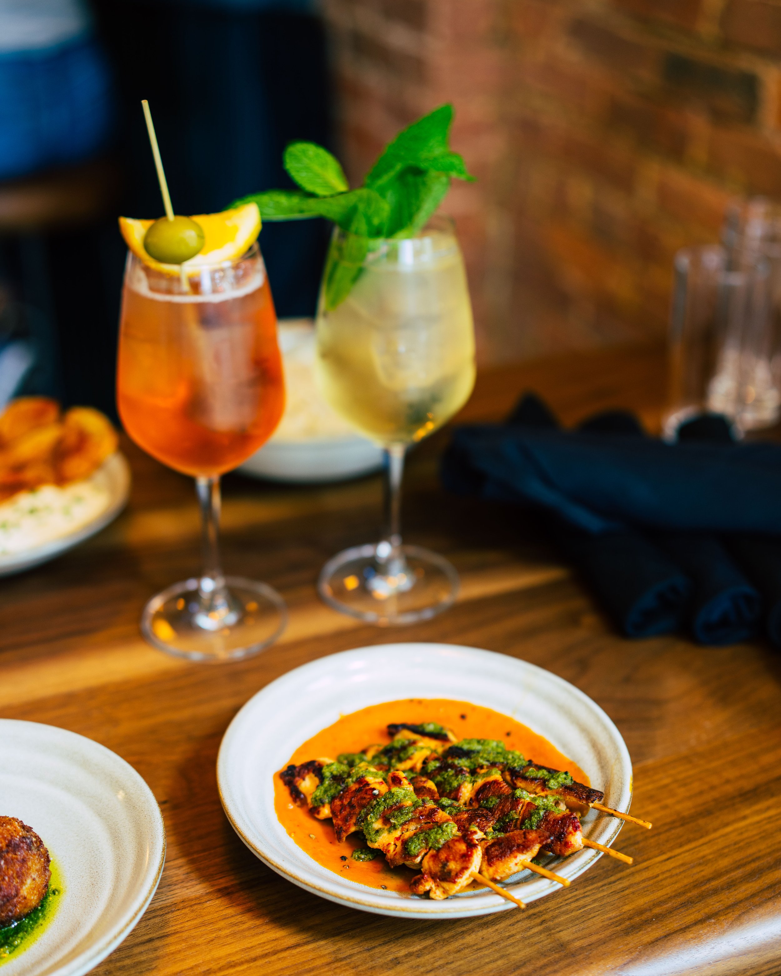 Two cocktails on a wooden table along with plates of food, including a plate of grilled chicken skewers.