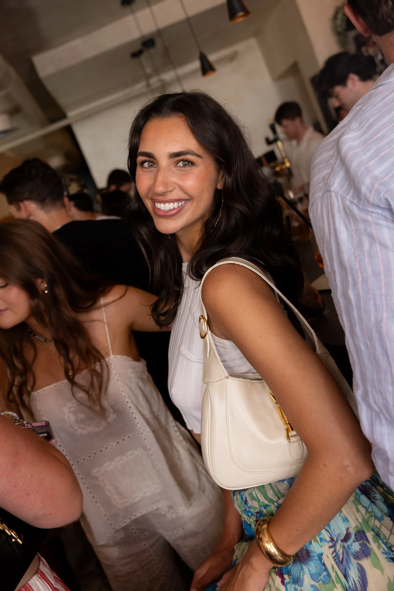 A young woman with long dark hair, smiling, in a sleeveless white top, carrying a white handbag, standing among a crowd at a social gathering in a warmly lit indoor space.