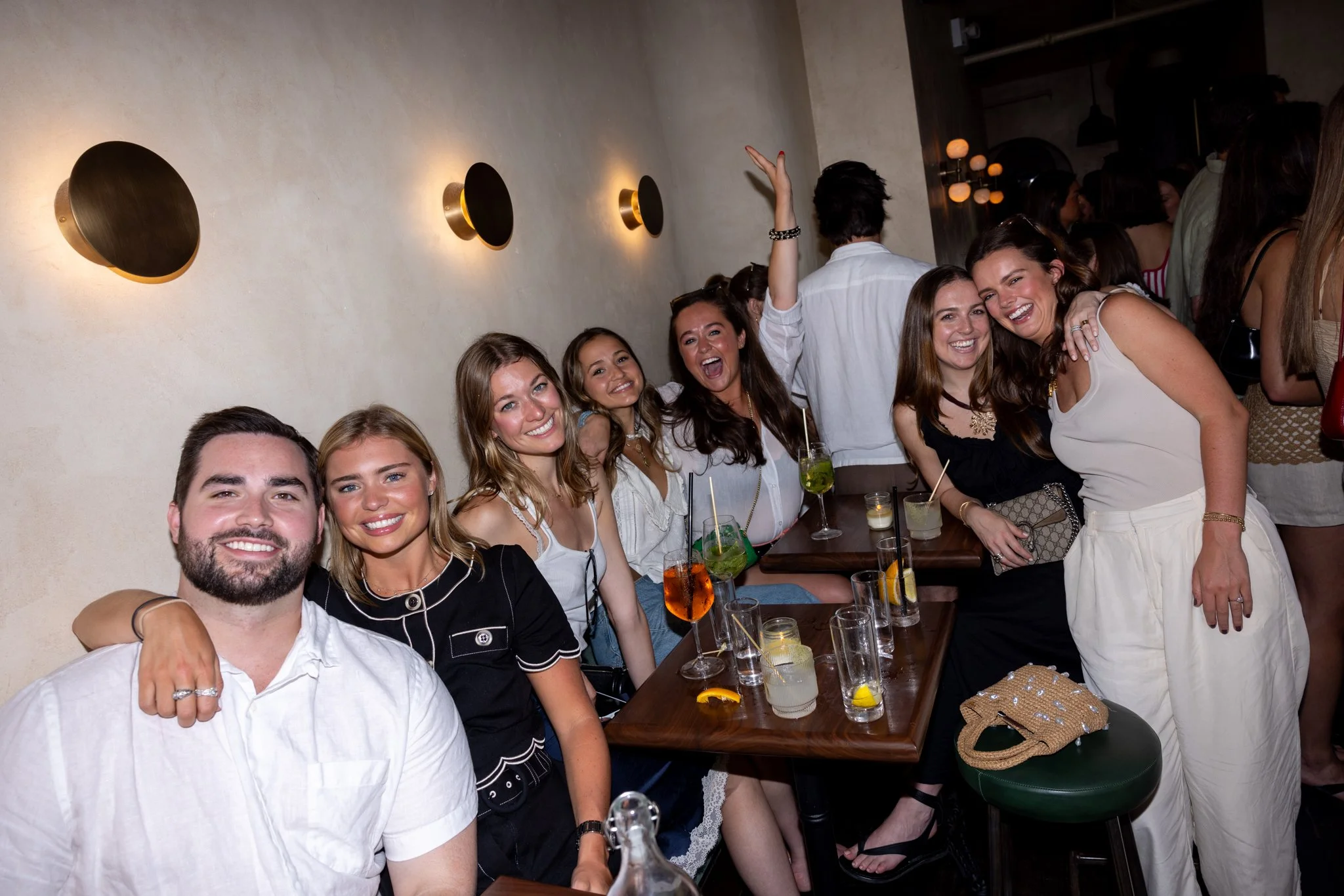 Group of smiling young adults enjoying drinks at a bar or restaurant, with some holding cocktails and others participating in a lively gathering.