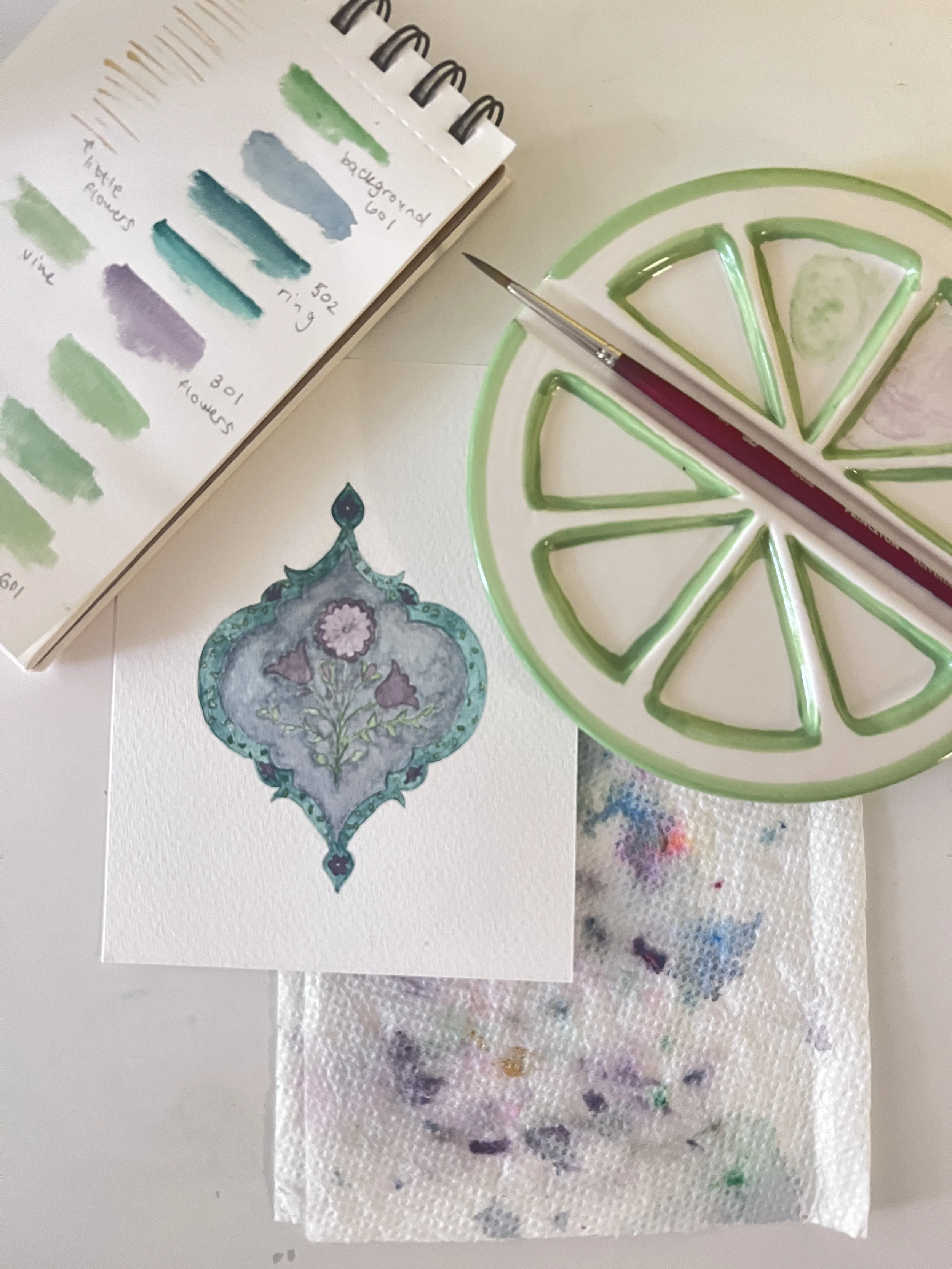 Watercolor paints and a palette with lime slices in green and pink, a paintbrush, a piece of paper with a blue and purple decorative floral design, a color swatch chart, and a paper towel with paint smudges.