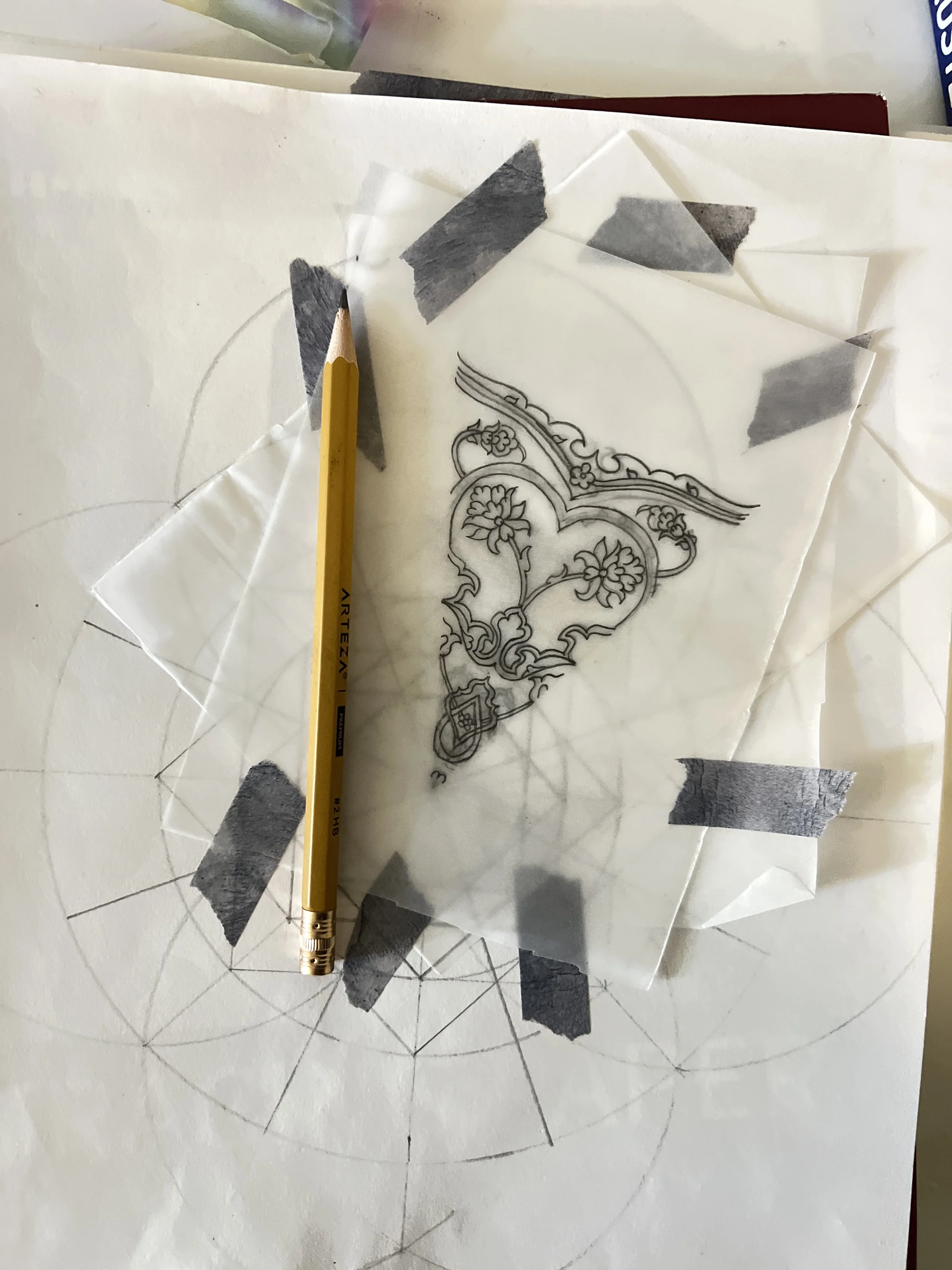 Sketch of intricate floral design on transparent paper, taped to larger circular pattern on white paper, with a yellow pencil resting on the tape.