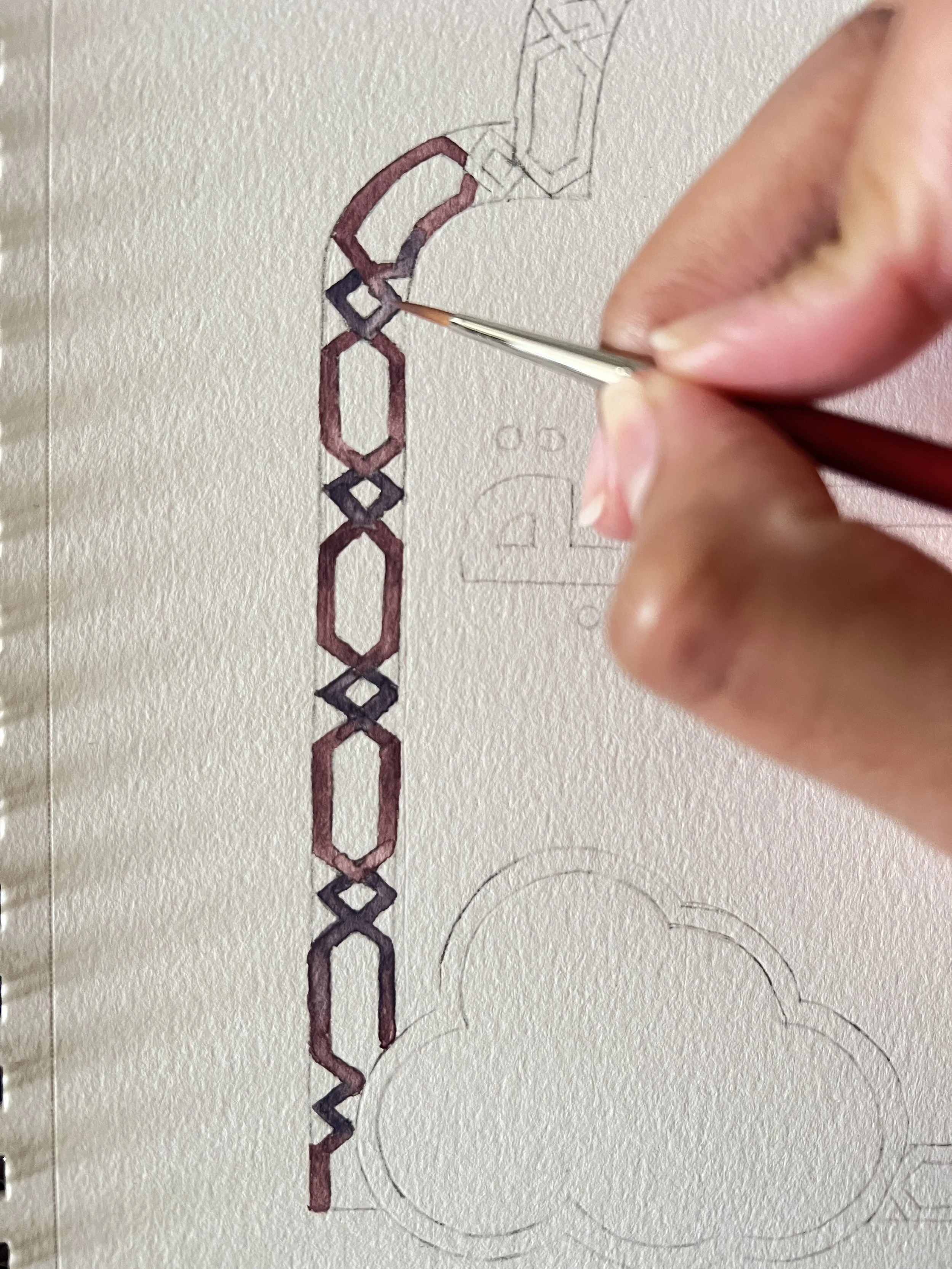 A person painting a chain-link pattern on paper with a small brush, with faint sketches in the background.