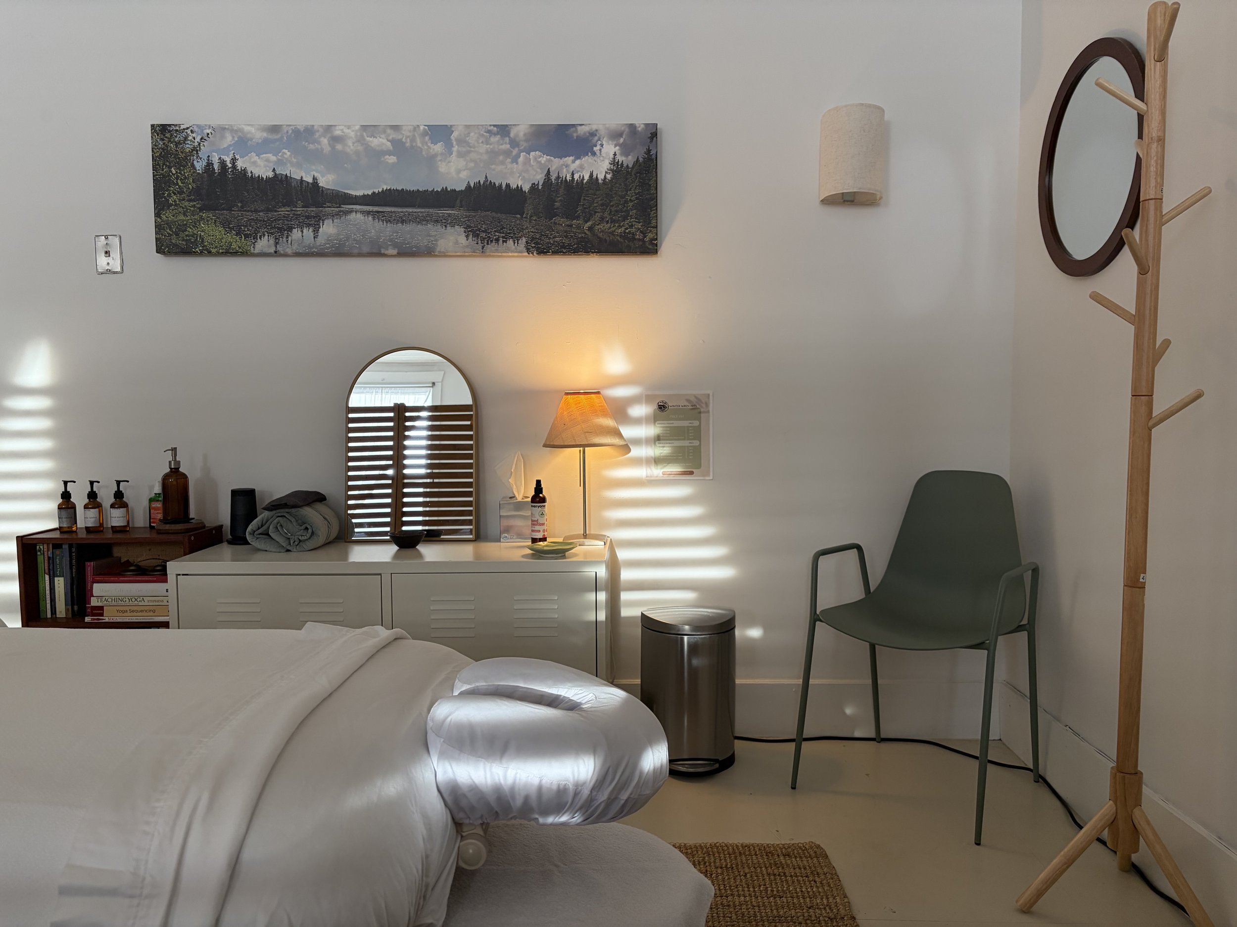 A photograph of a bright white room with a dressed massage table and a green chair in the corner