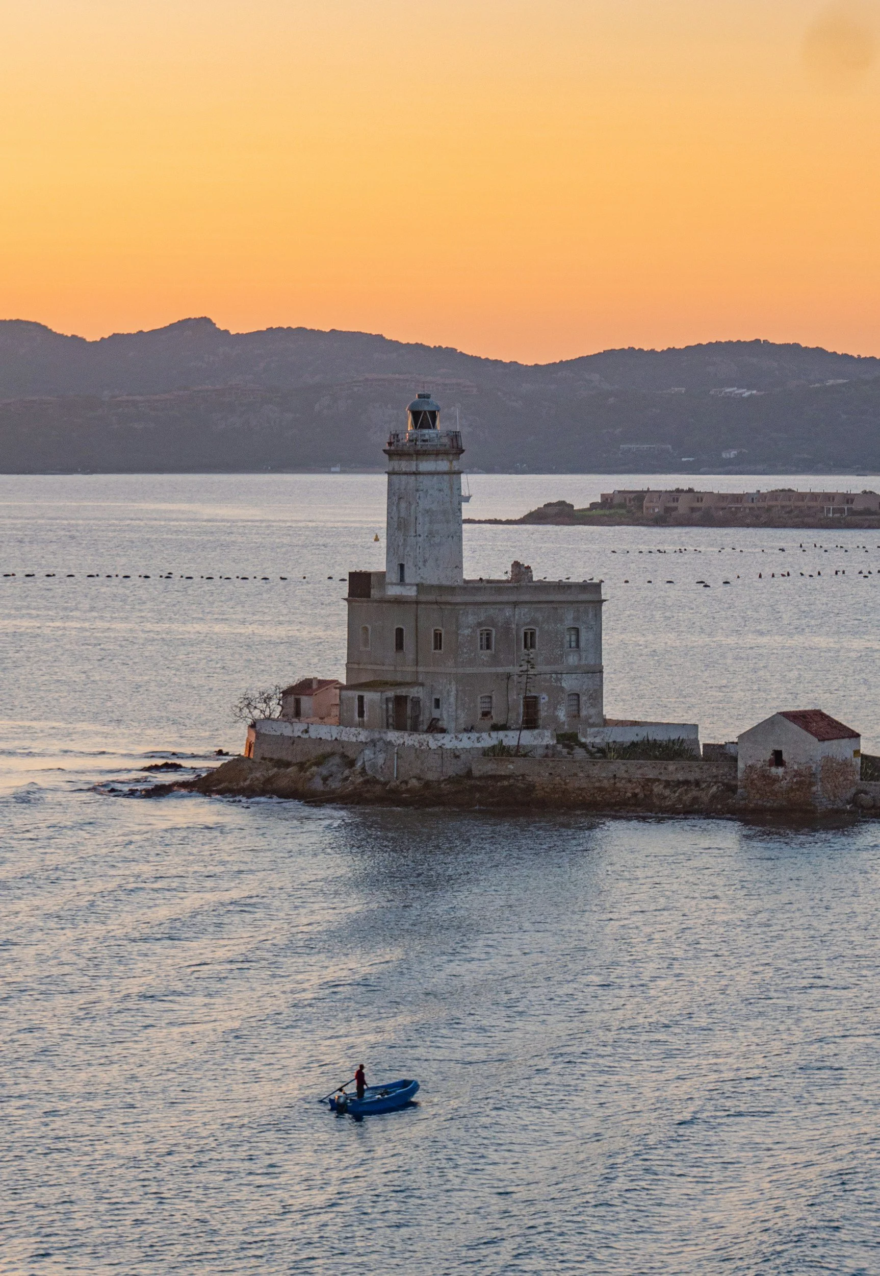 A lighthouse on a small island with a building at its base, water surrounding the island, a person on a small boat below, and mountains in the background at sunset.