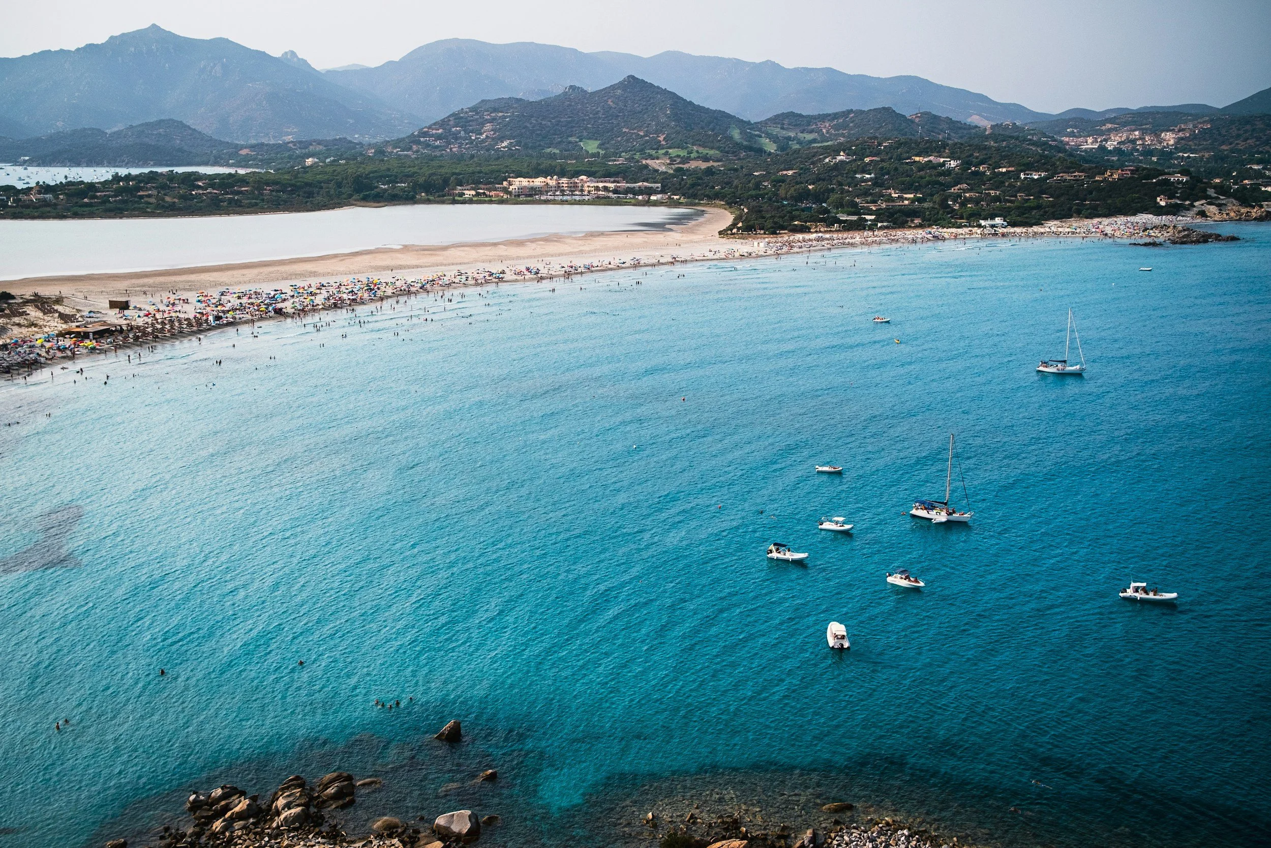 A coastal scene with a sandy beach filled with colorful umbrellas and beachgoers, turquoise water with several boats, rocky shoreline in the foreground, and green hills and mountains in the background under an overcast sky.