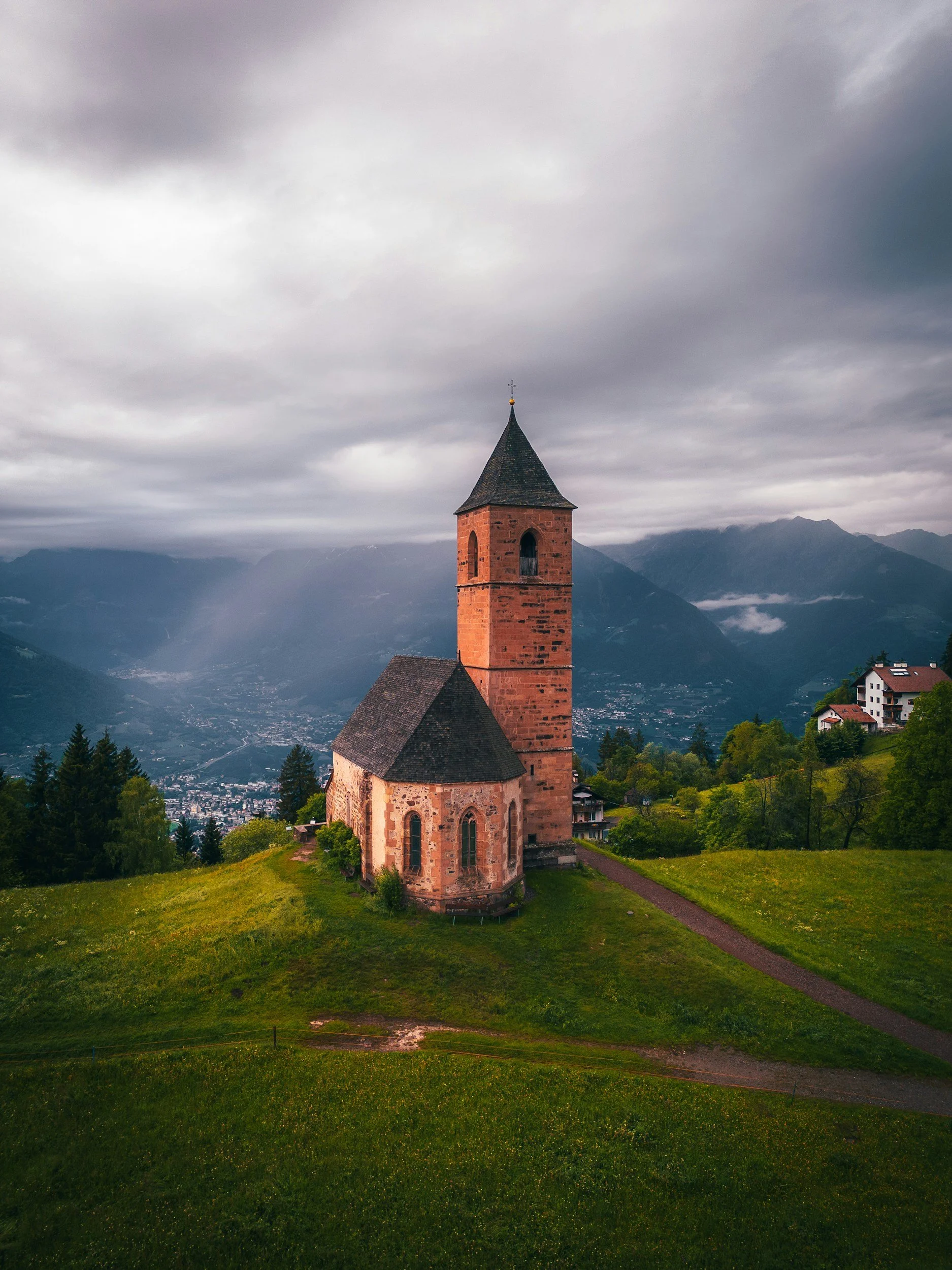 A church with a tall brick steeple on a grassy hill, surrounded by trees, mountains, and cloudy skies in the background.