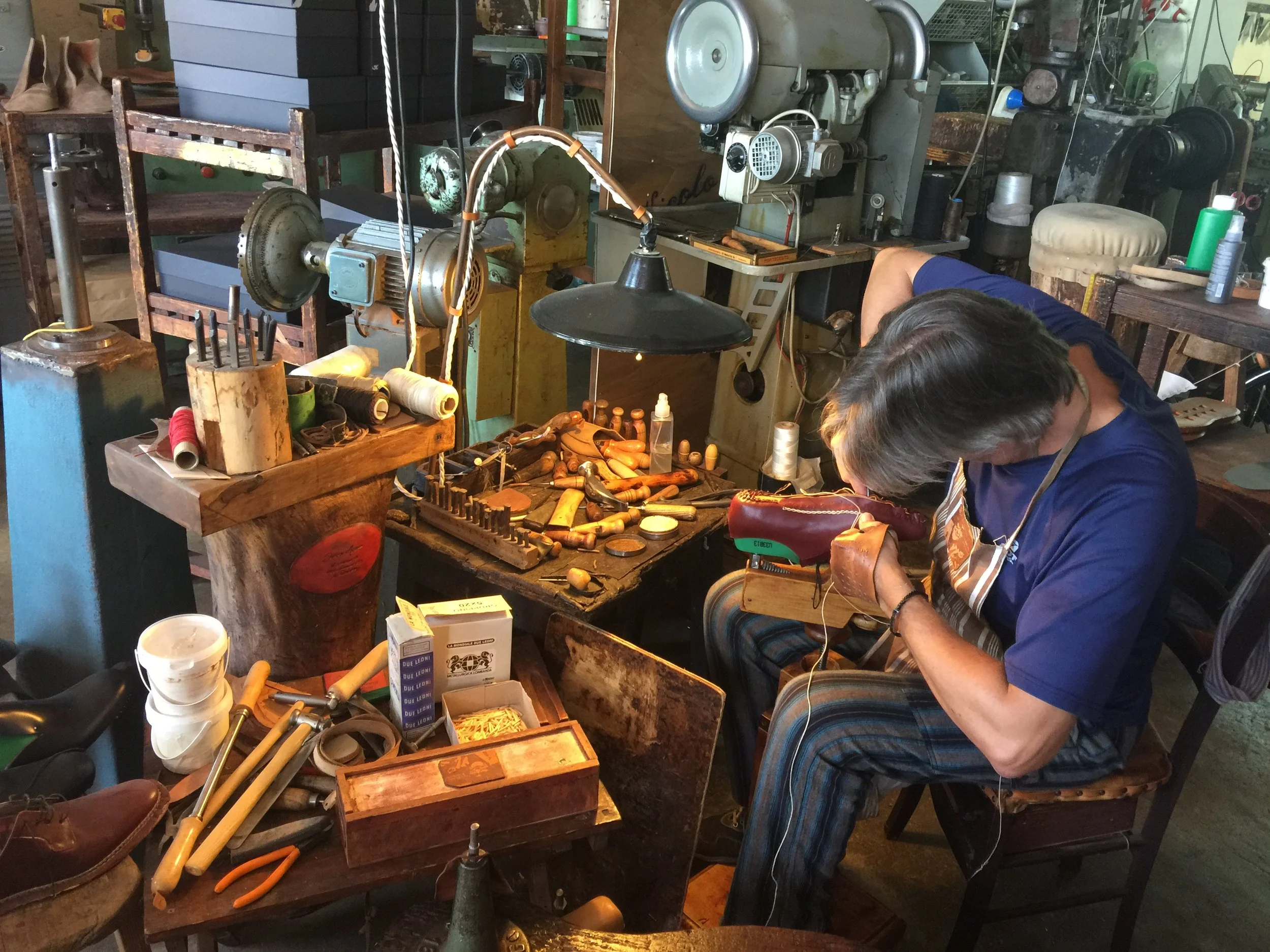 A person working on a leather item in a leather workshop, surrounded by tools, spools of thread, and leatherworking equipment.