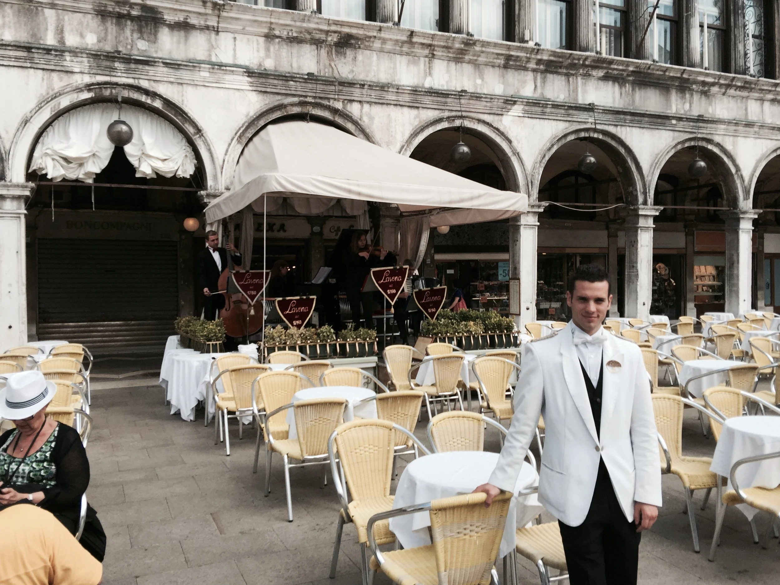 Man in white tuxedo standing at an outdoor cafe, with live music performers in the background.