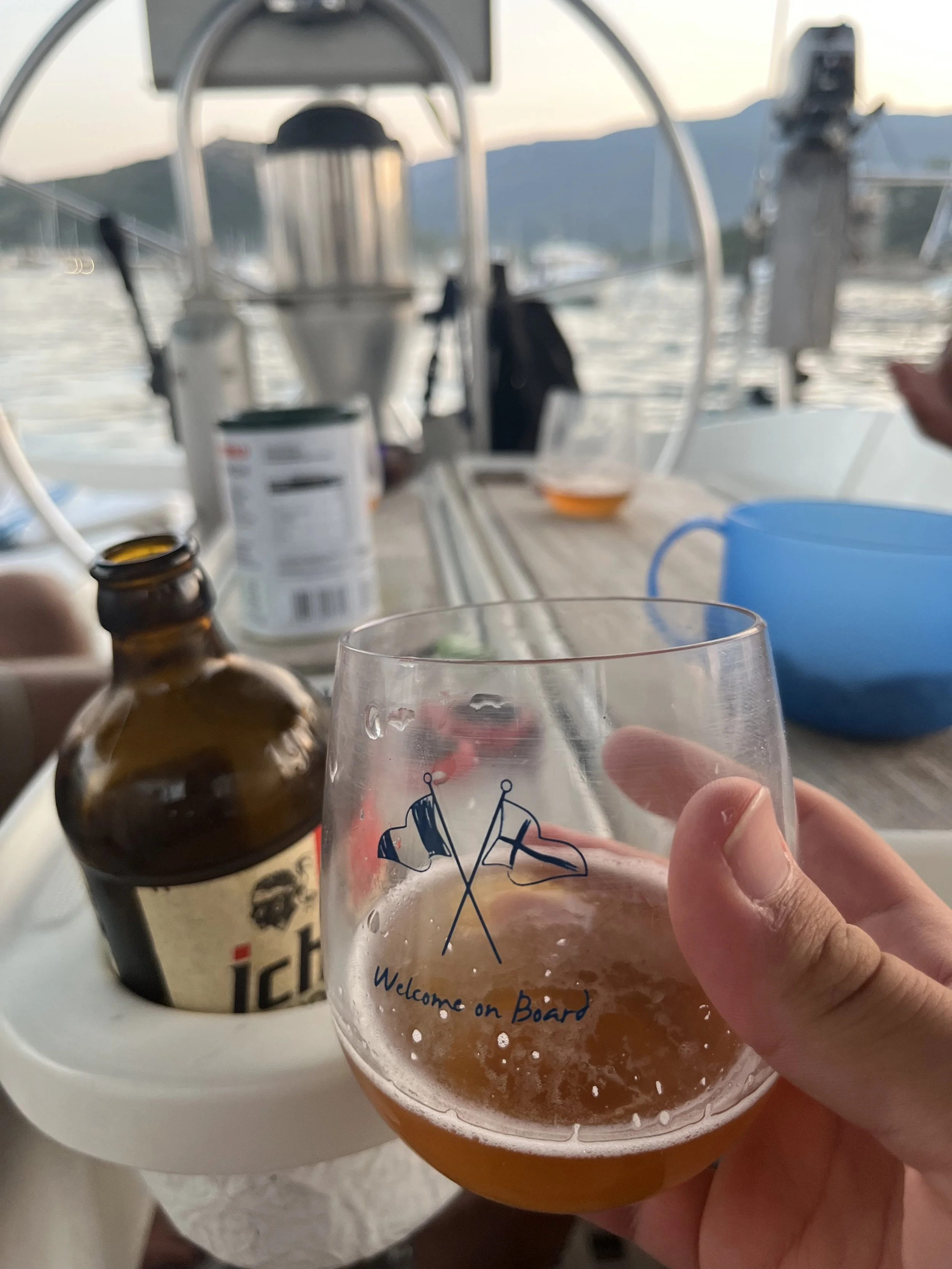A hand holding a glass with a beer on a boat, with a bottle of beer on a tray behind it, and a table with various items, with water and mountains in the background.