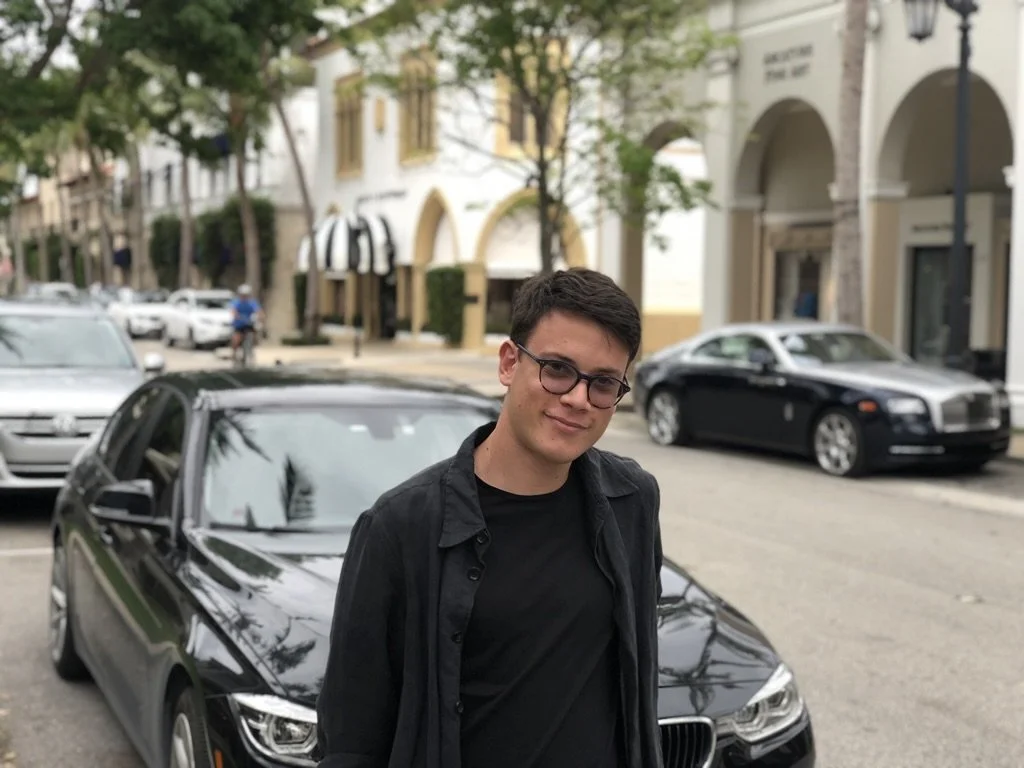A young man with short dark hair and glasses stands in front of a black BMW parked on a city street, with buildings and more cars in the background.