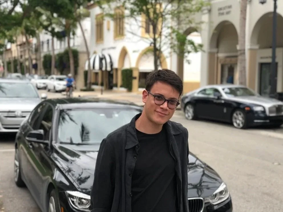 A young man wearing glasses and a black shirt standing in front of a black car on a city street with trees and buildings.