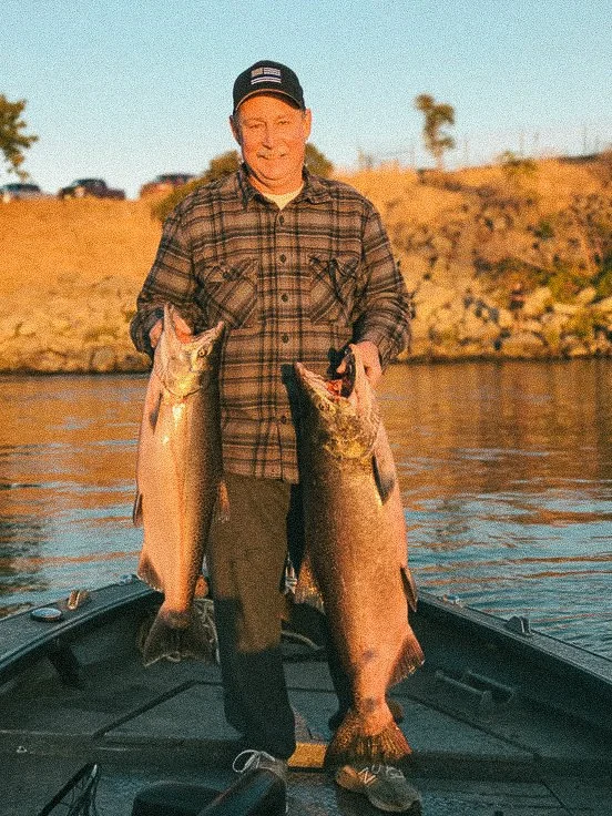 A man standing on a boat holding two large fish, one in each hand, with a river and rocky shoreline in the background during sunset.