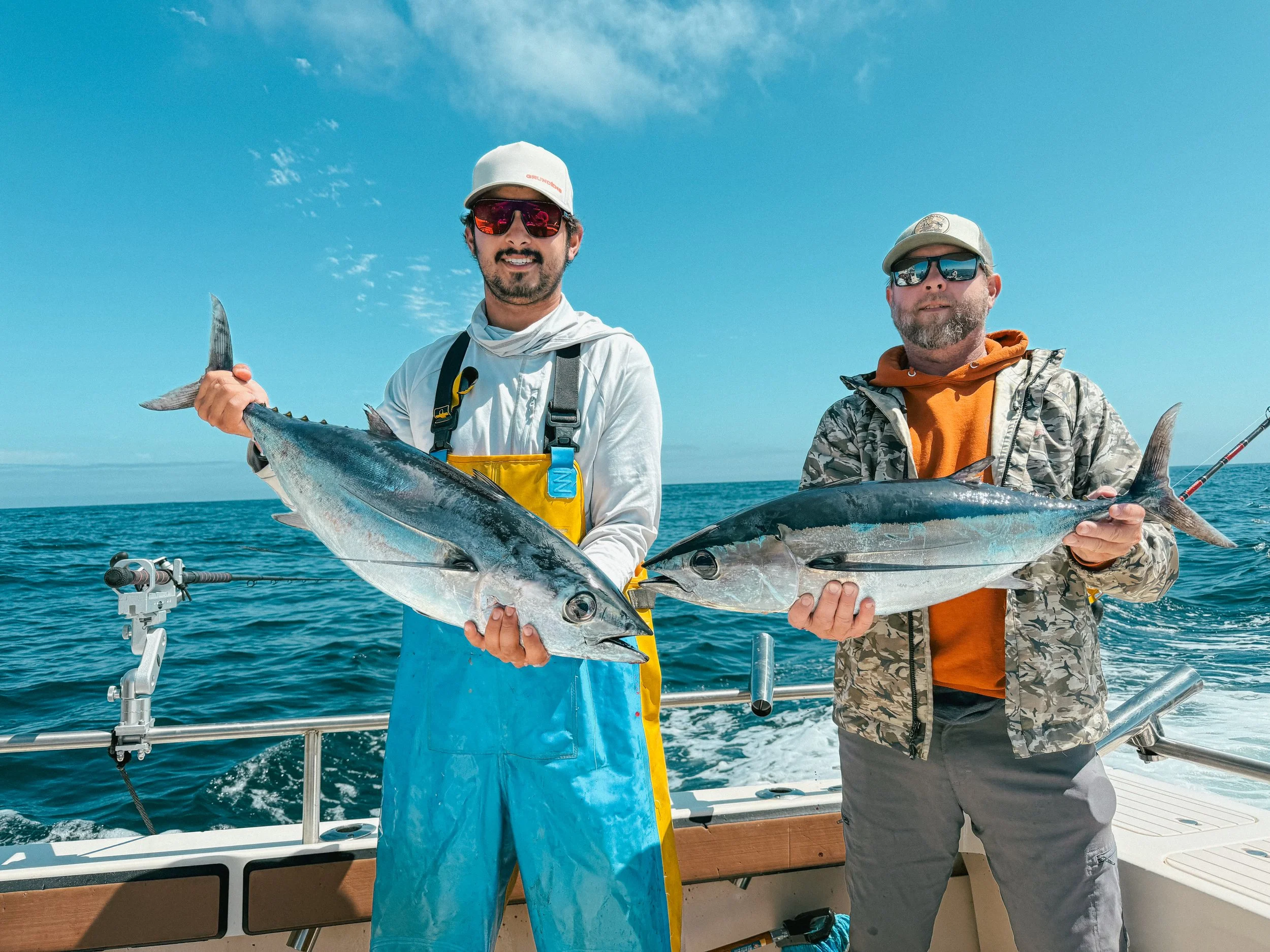 Two men on a boat holding caught fish with the ocean and blue sky in the background.