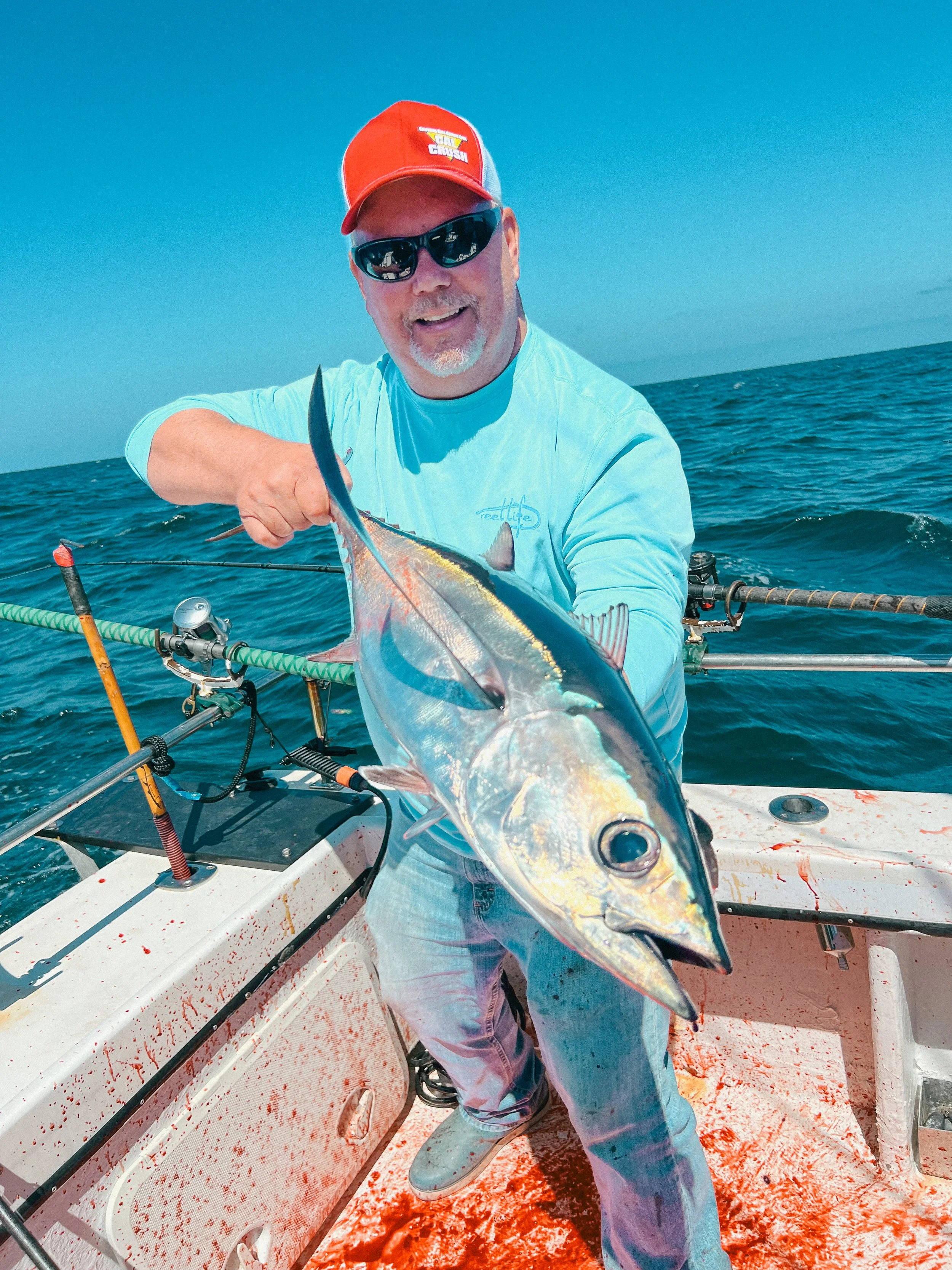 A man wearing a red cap, sunglasses, and a light blue shirt is holding a large fish on a boat in the ocean.