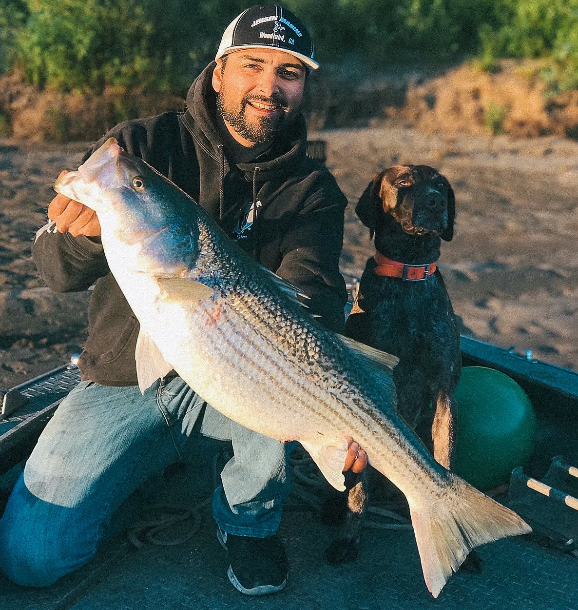 Man holding a large fish with a dog sitting next to him on a boat, outdoors near a river or lake.