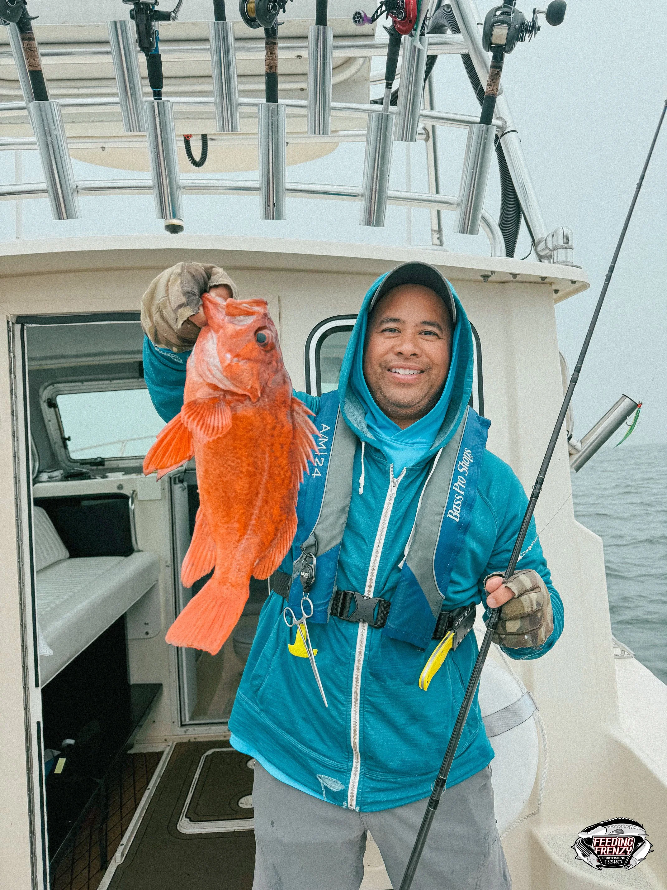 A man in blue jacket and gloves holding a large orange fish on a boat, smiling. The boat has rods and fishing gear, and the open doorway shows the boat's interior. There's a logo on the bottom right corner for Feeding Frenzy.