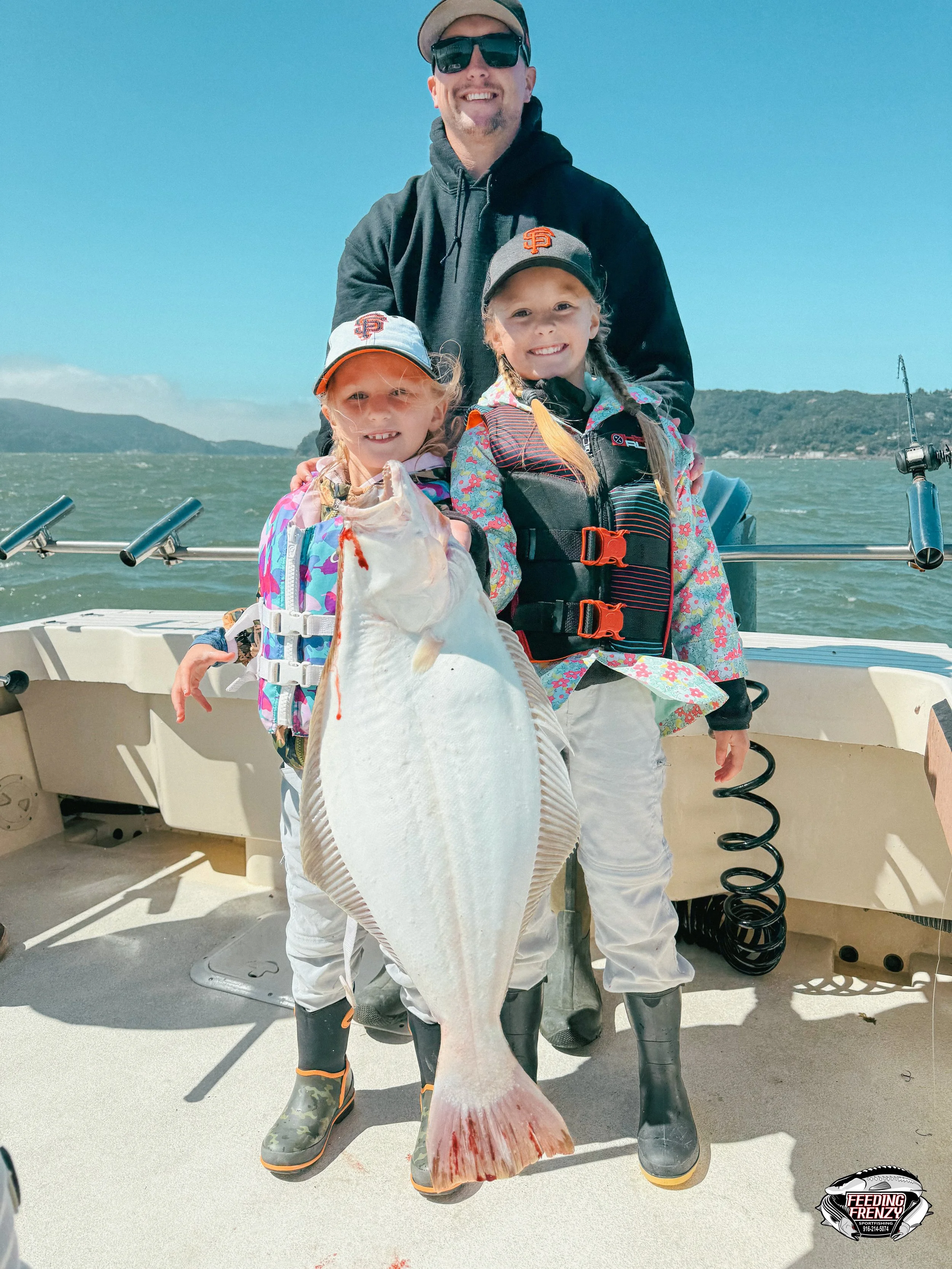 Two young girls and an adult male fishing on a boat. The girls are holding a large fish. The man is standing behind them, smiling, in a black jacket and sunglasses. The girls are wearing colorful jackets, hats, and boots. The boat is on a body of wat