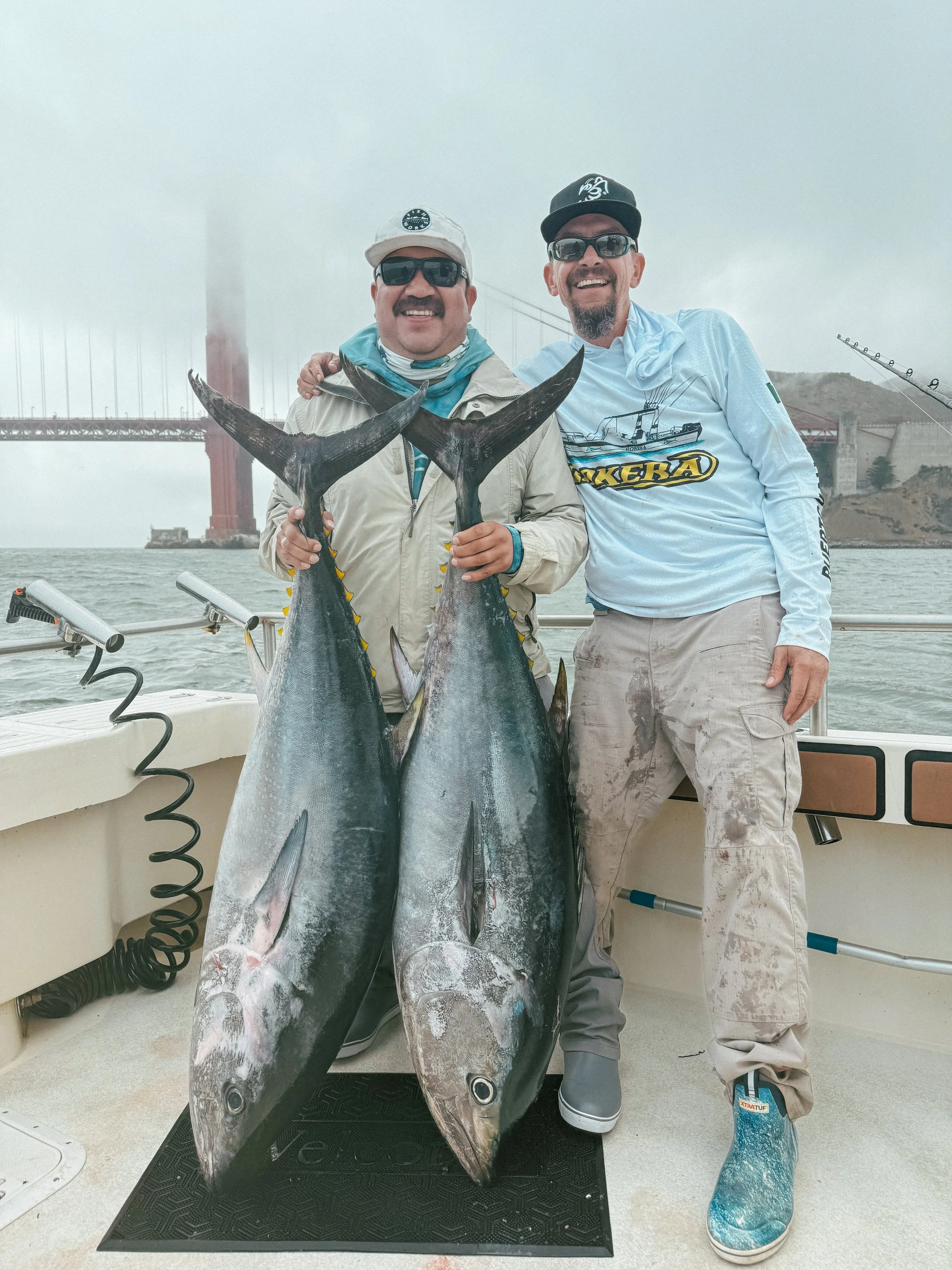 Two men on a boat holding large fish, with the Golden Gate Bridge in the background.