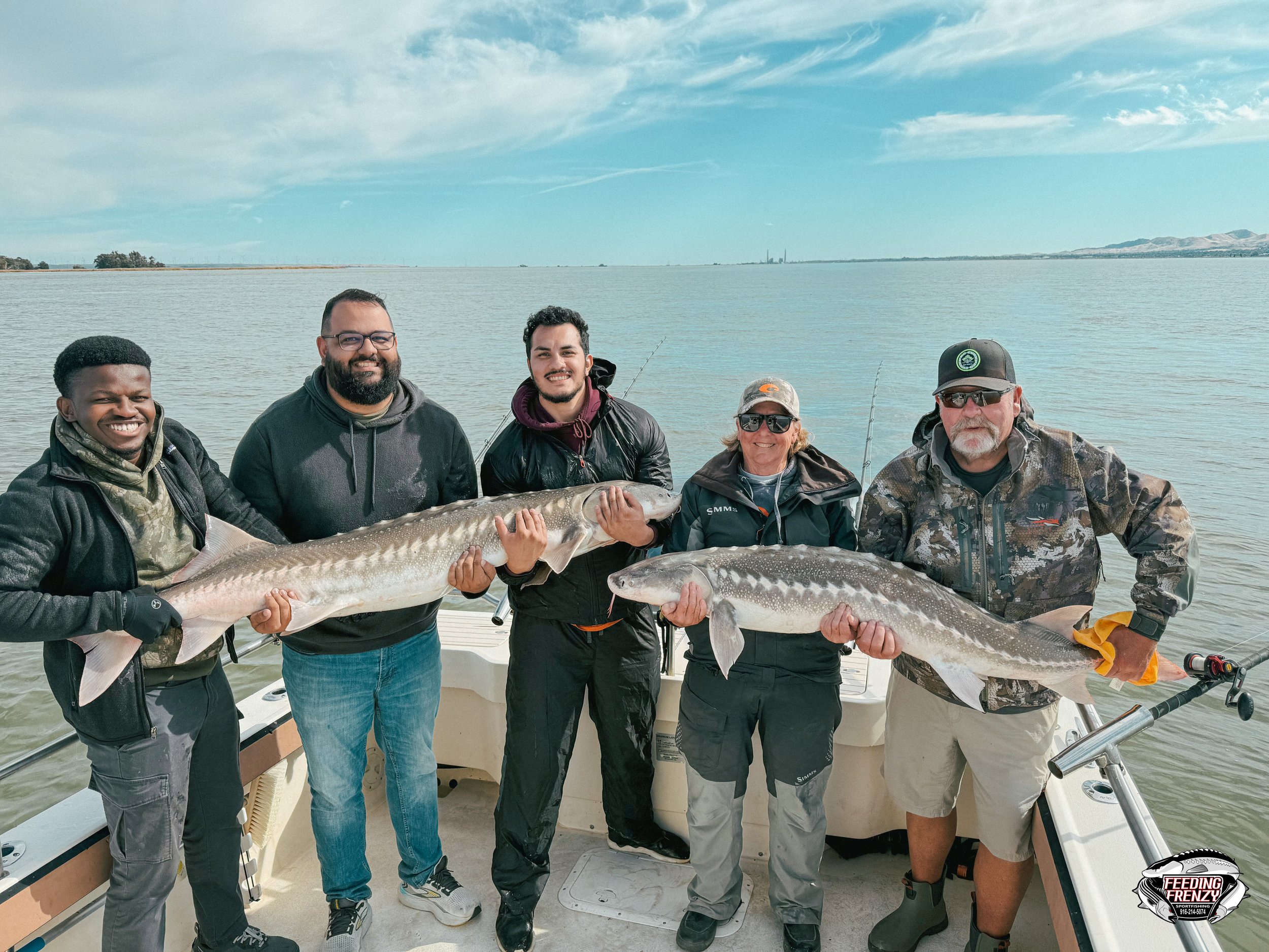 Group of five people on a boat holding two large fish, with a body of water and distant shoreline in the background.