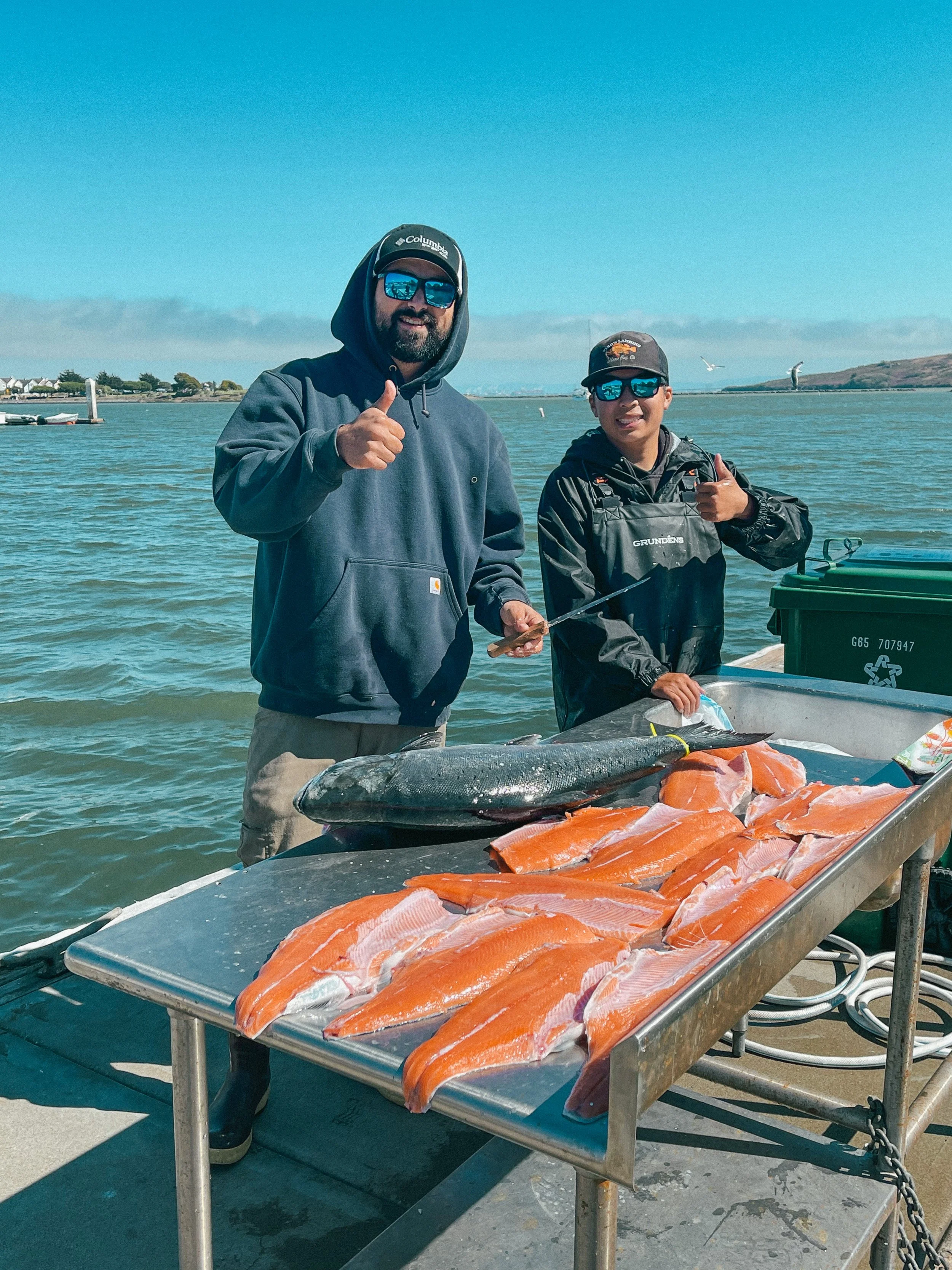 Two men standing on a dock next to a table with freshly caught fish, including salmon and a large fish, with water and boats in the background.