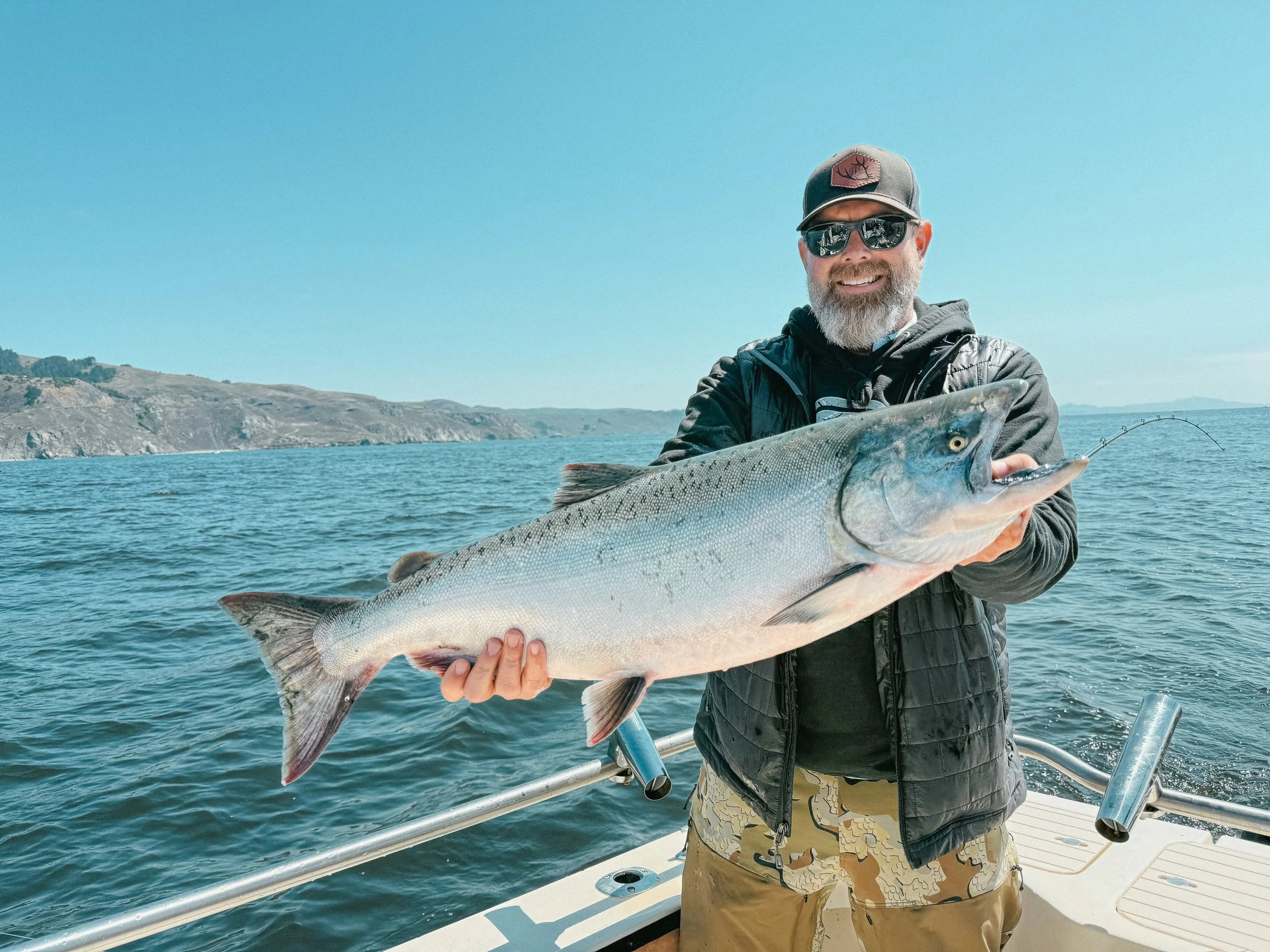 A man with sunglasses and a beard holding a large fish on a boat in the water, with scenic hills in the background.