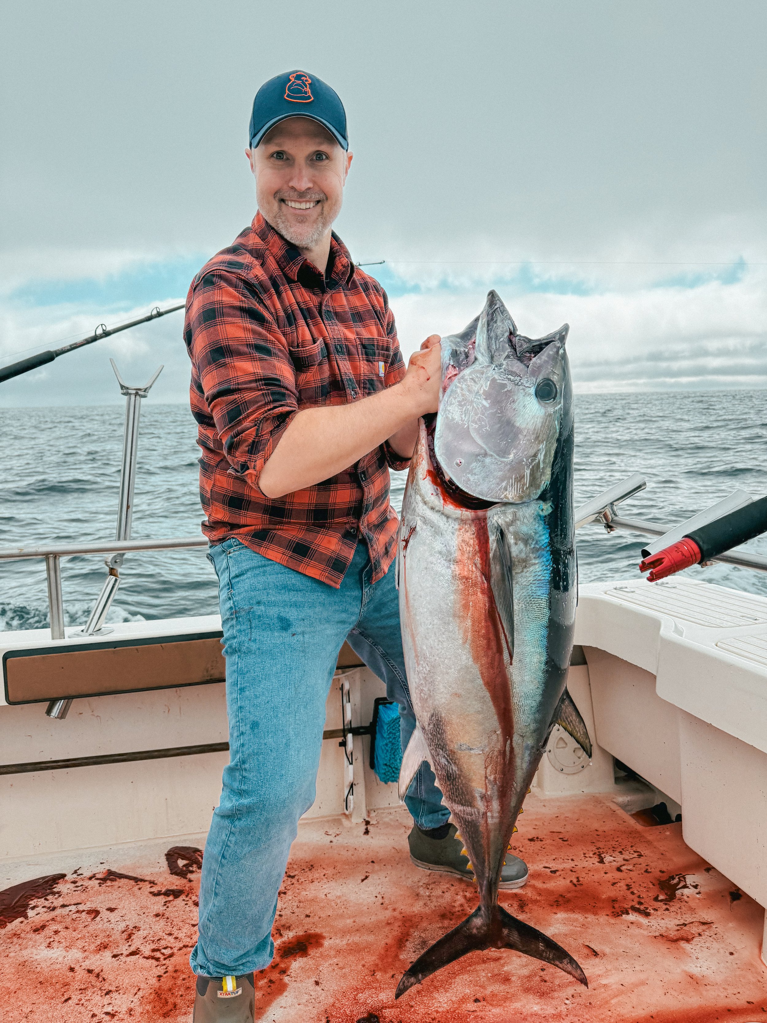 A man in a plaid shirt and baseball cap holding a large fish on a boat at sea.