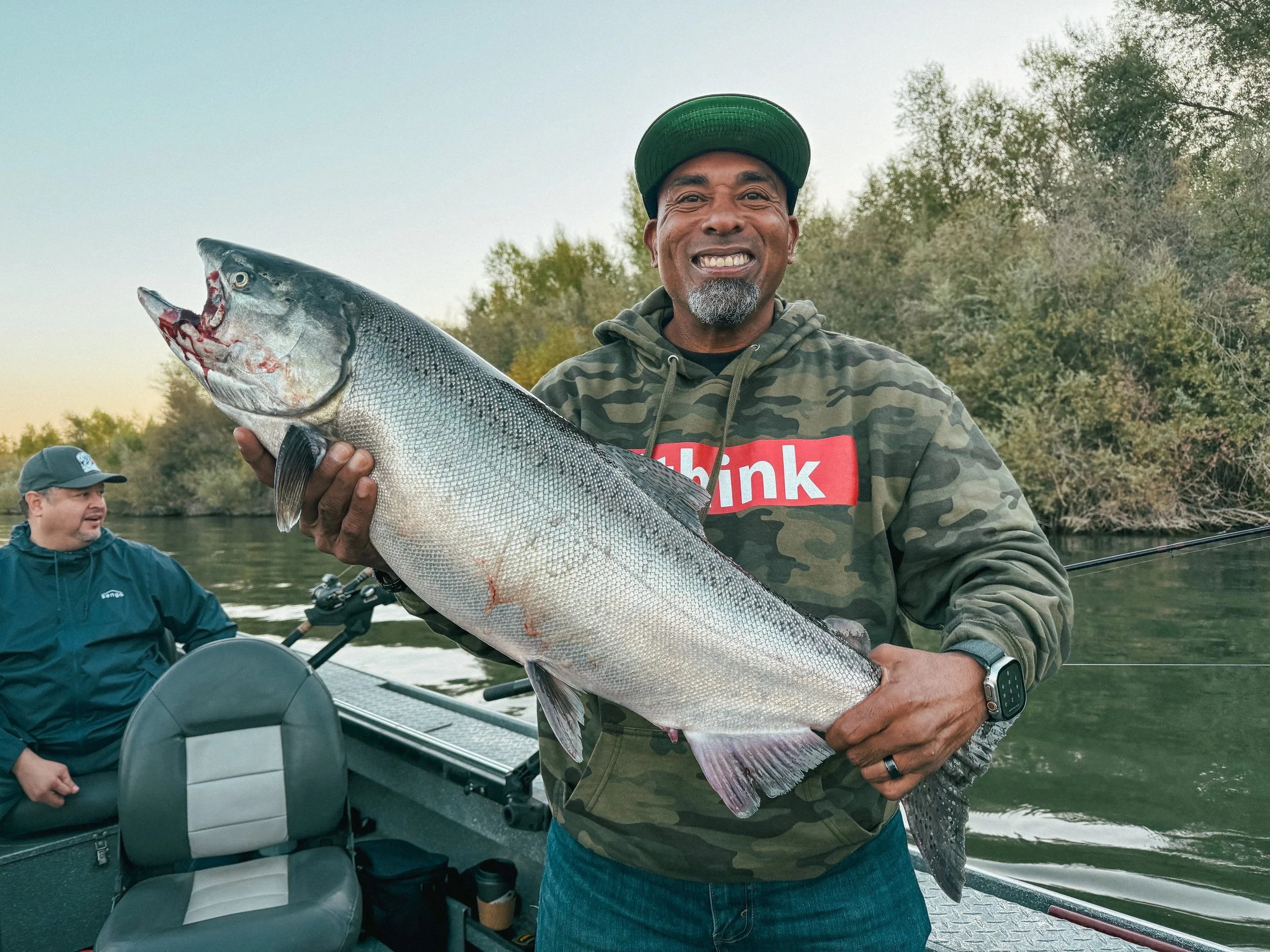 A man smiling and holding a large fish he caught on a boat during a fishing trip, with another man sitting in the background.