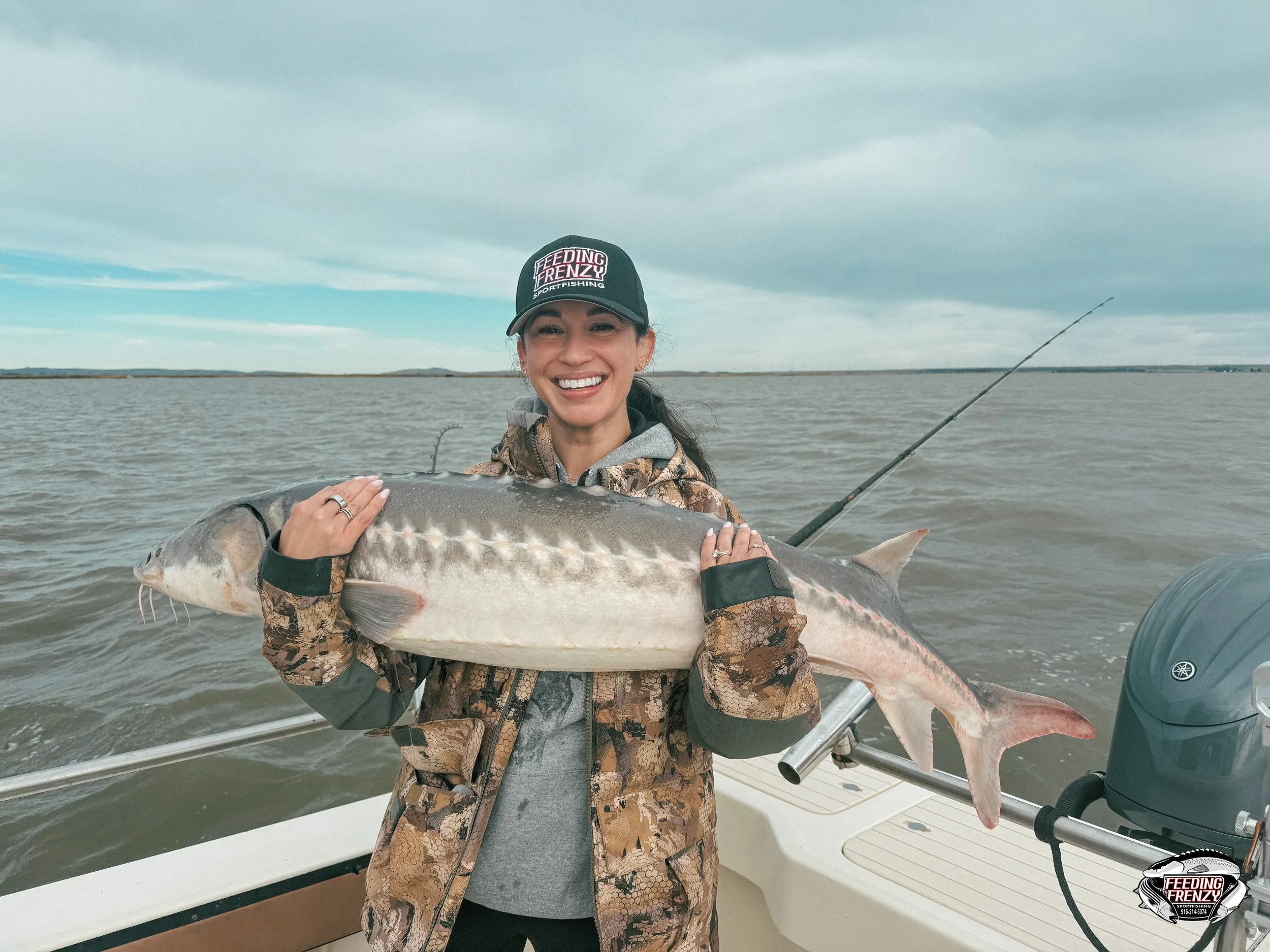 A woman holding a large fish on a boat, smiling, with water and cloudy sky in the background.