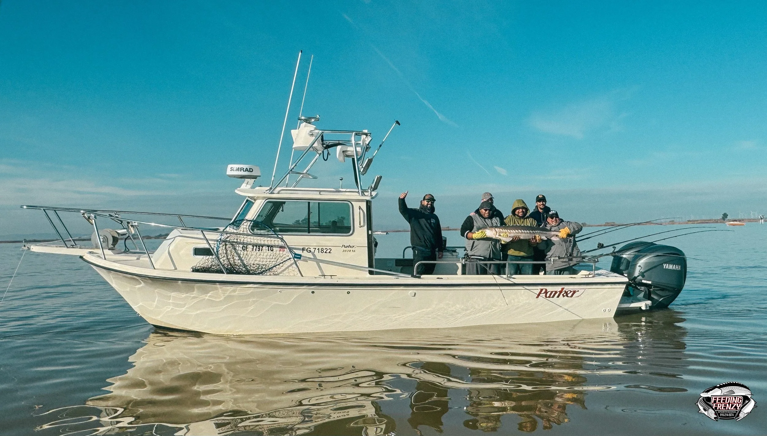Six people on a boat holding a large fish, with water and a clear sky in the background.