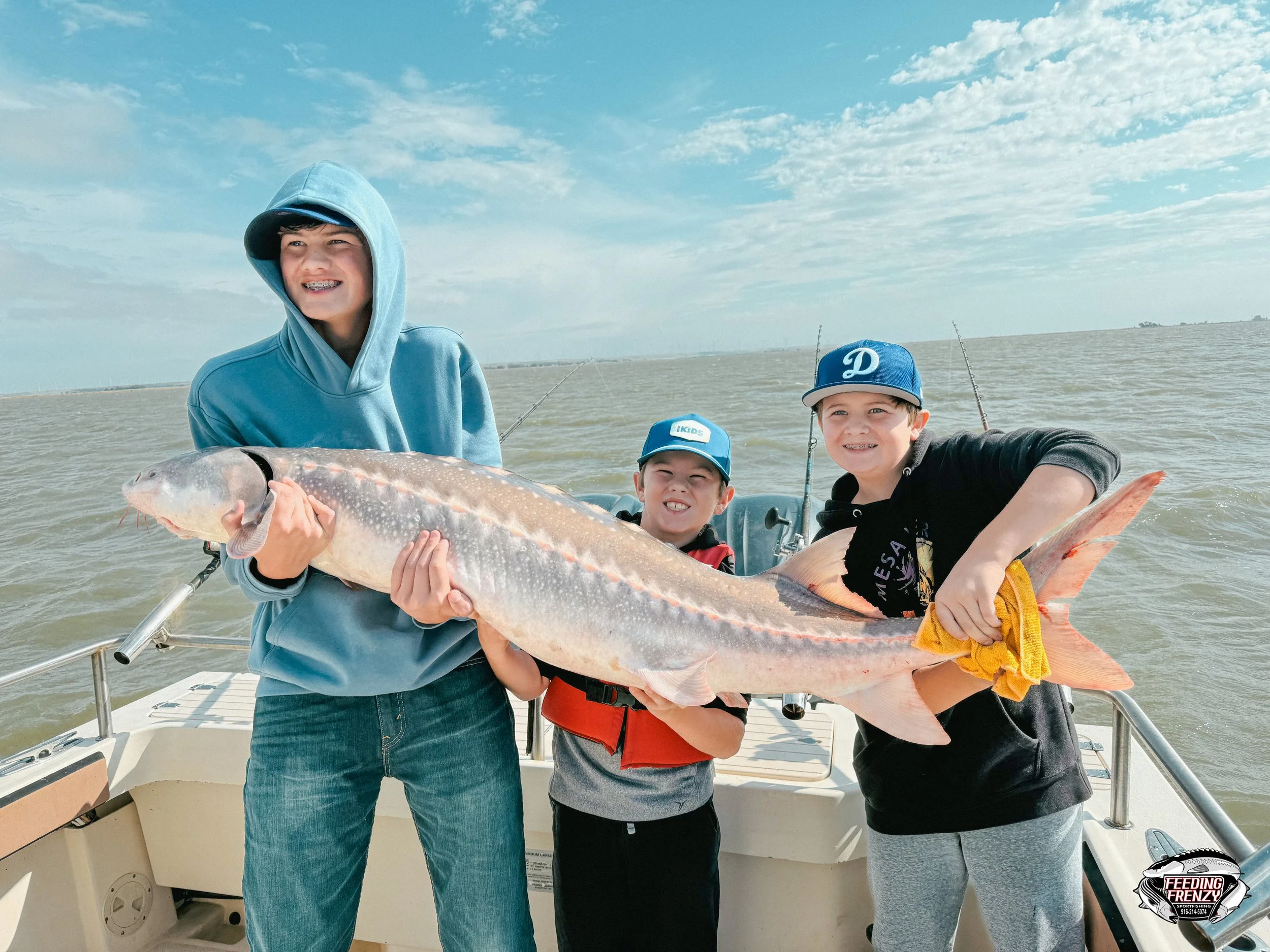Three boys on a boat holding a large fish they caught during a fishing trip. They are smiling and wearing casual clothing and hats. The background shows water and a partly cloudy sky.
