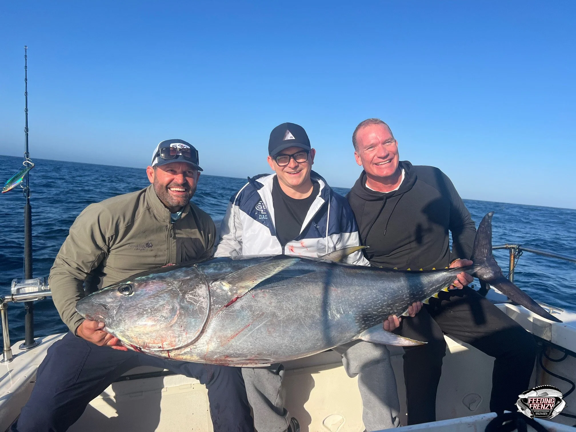 Three men on a boat holding a large fish they caught, with the ocean and clear blue sky in the background, celebrating their fishing success.