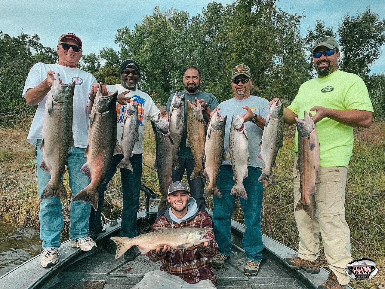 Six men holding large fish, standing on a boat, with one man kneeling in front holding a fish, in a natural outdoor setting with trees and grass.