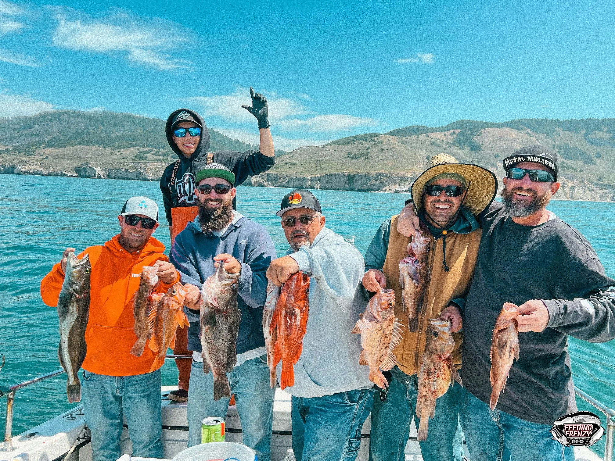 Group of seven men on a boat holding freshly caught fish, with scenic water and hills in the background, sunny weather, and some wearing hats and sunglasses.