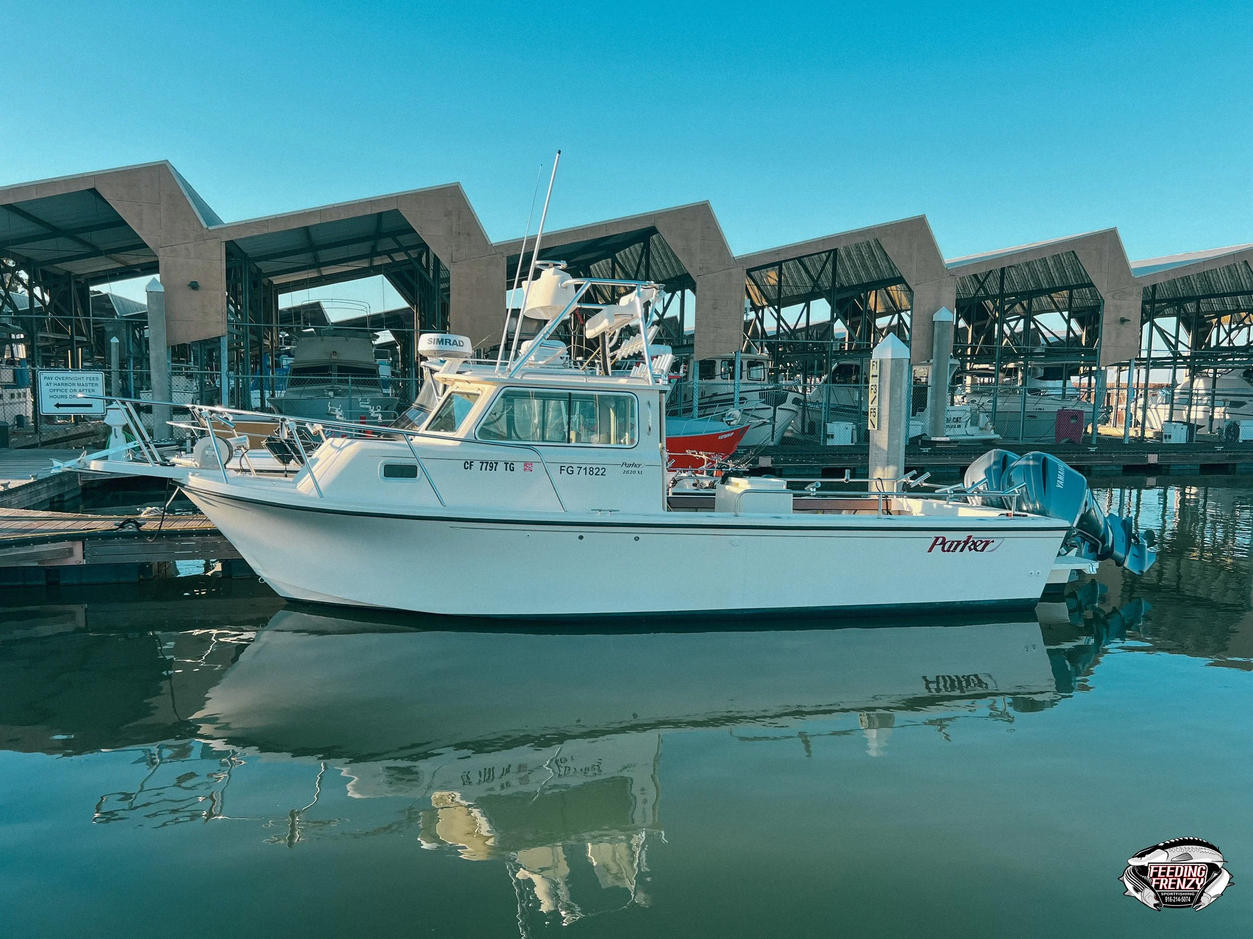 A white boat named 'Parker' docked at a marina with other boats and a modern covered boat storage structure in the background under a clear blue sky.