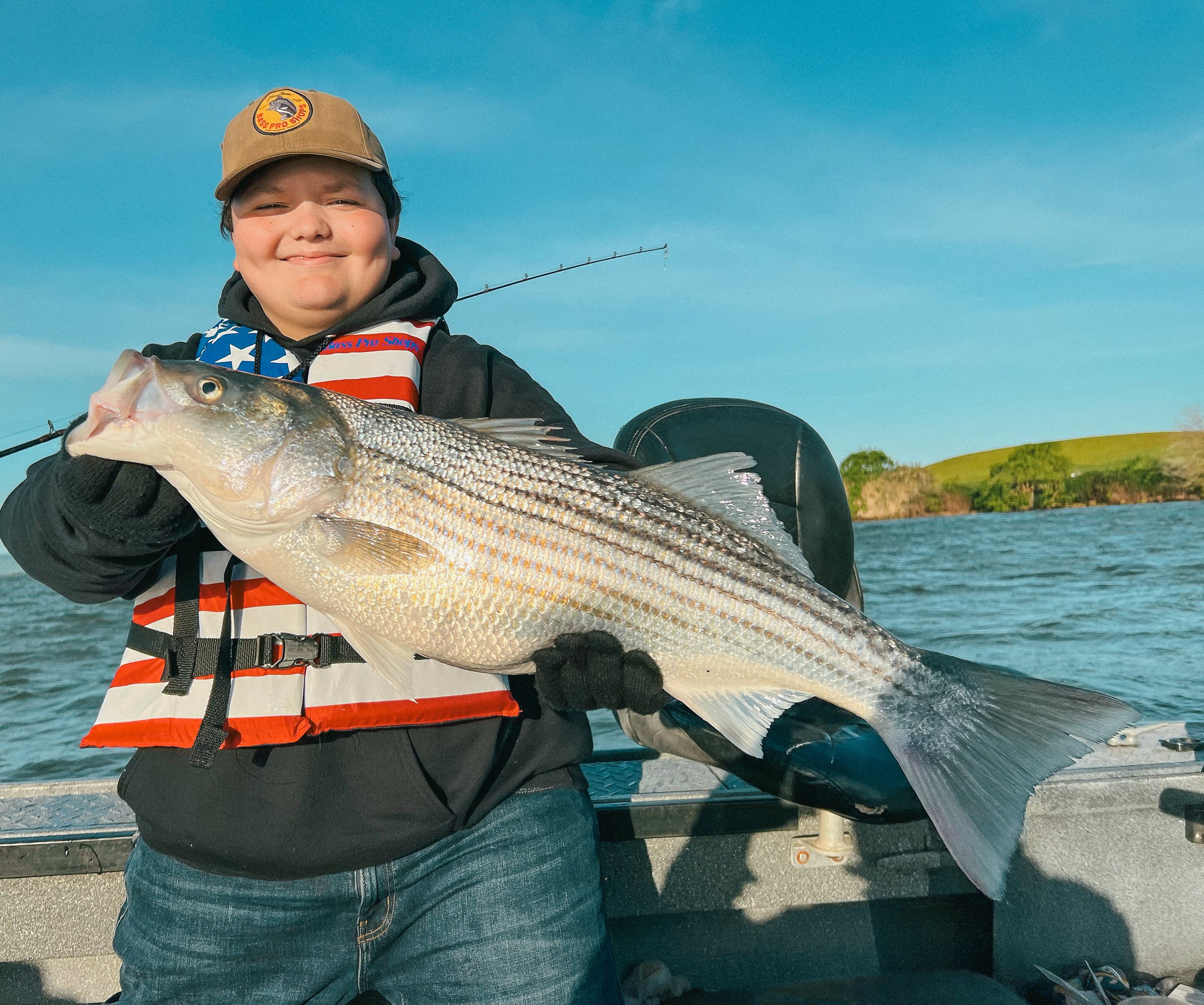 A boy smiling and holding a large striped bass fish on a boat with water and green landscape in the background.