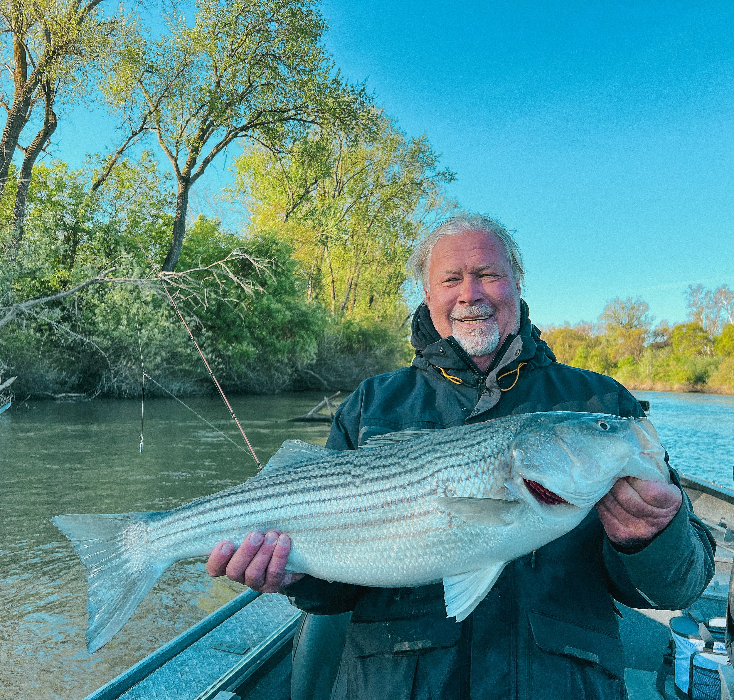 An older man smiling and holding a large fish on a boat, with a river and green trees in the background on a clear day.