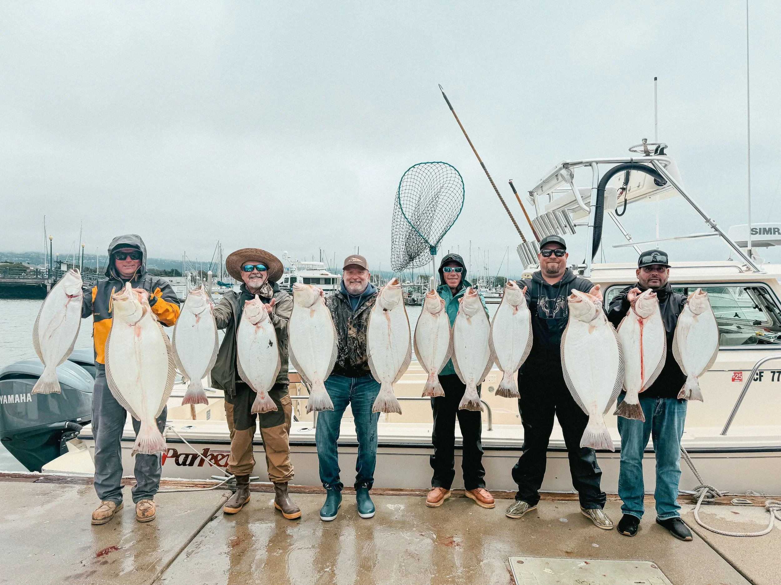 Six men standing on a dock in front of a boat, each holding a large flatfish, with a marina and cloudy sky in the background.