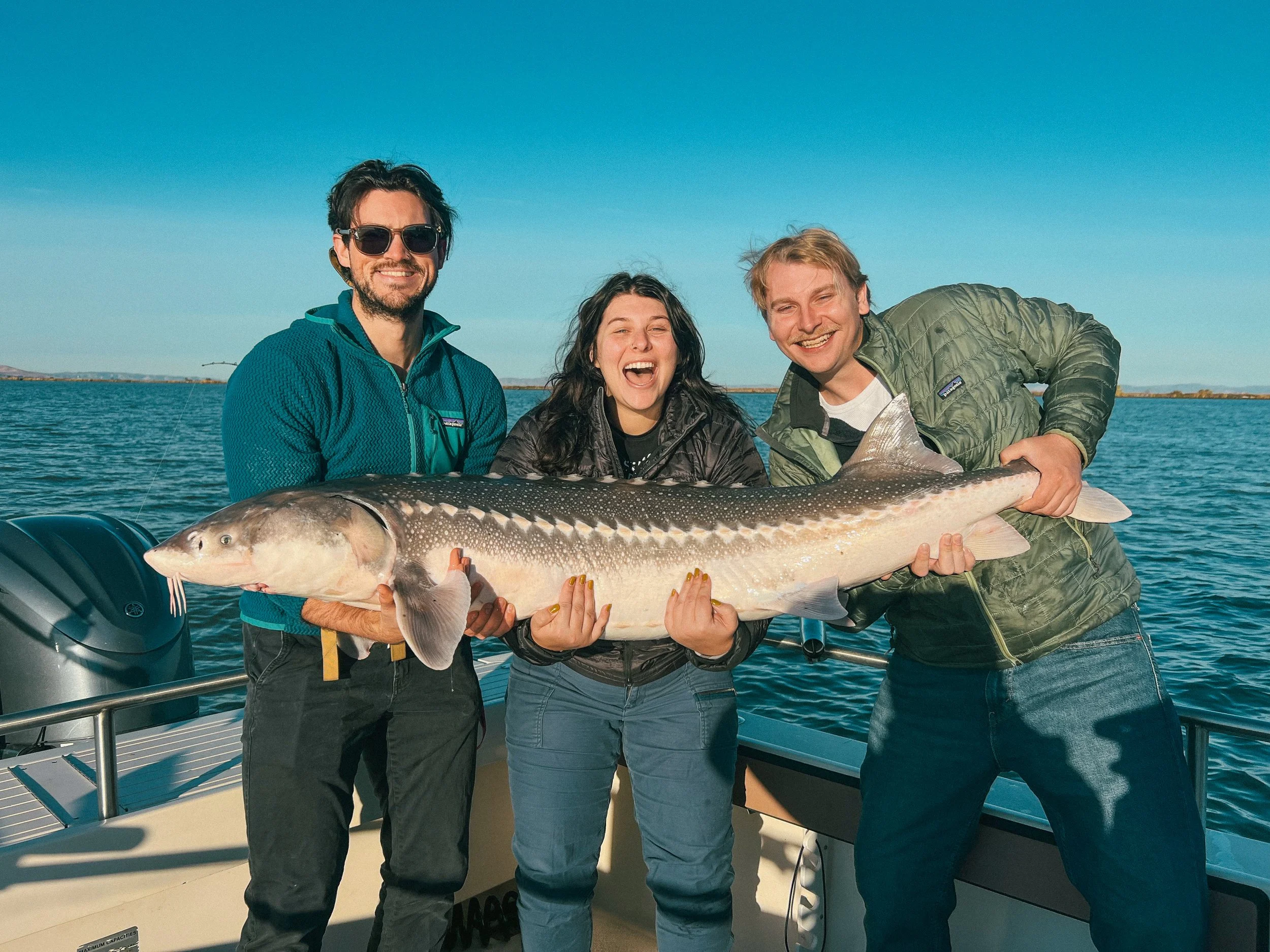 Three people on a boat holding a large fish, smiling, with water and sky in the background.