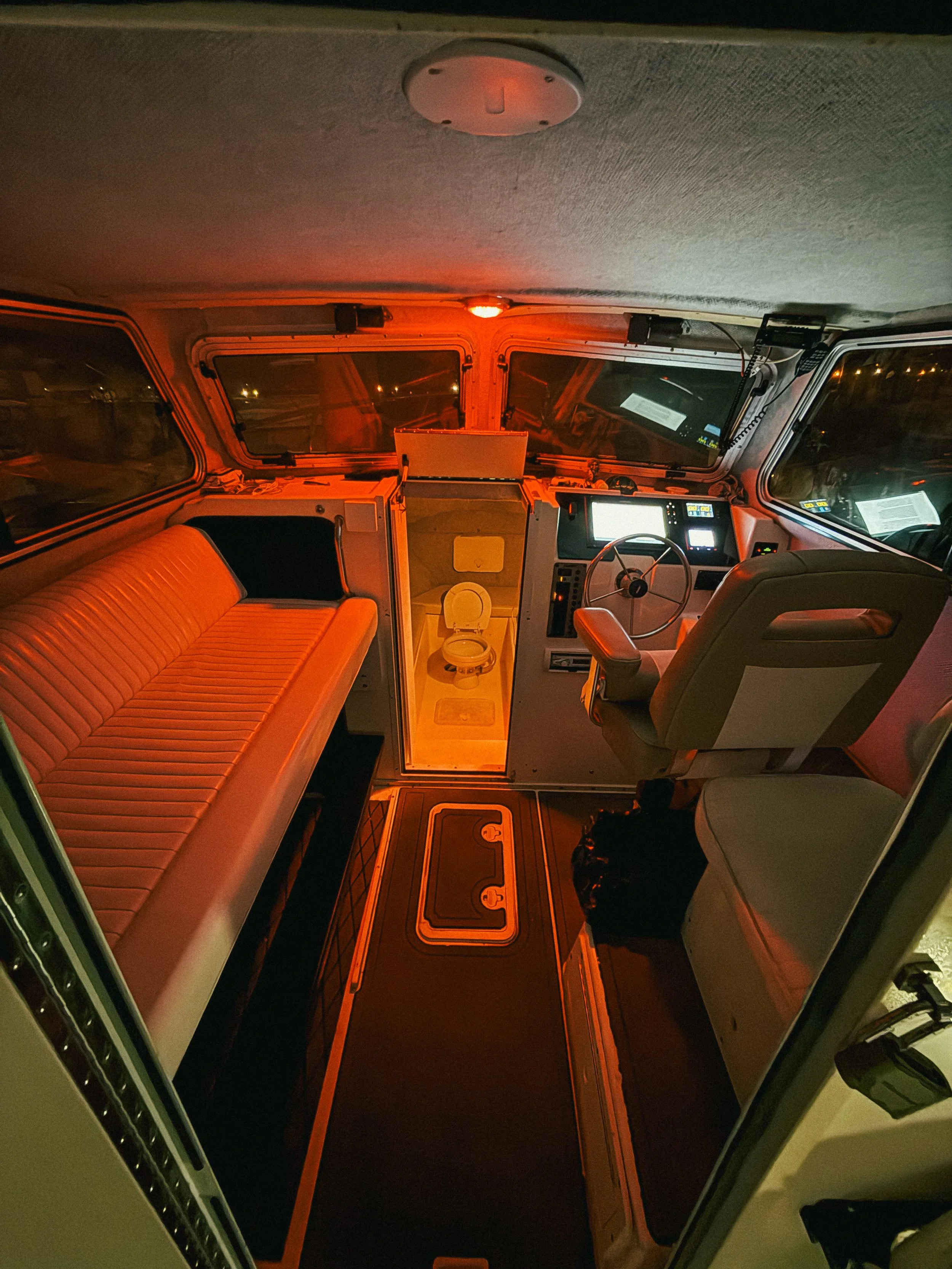 Inside of an RV or bus at night, showing a front seat and a bench seat, with a small bathroom between them, illuminated by orange and some white lights.