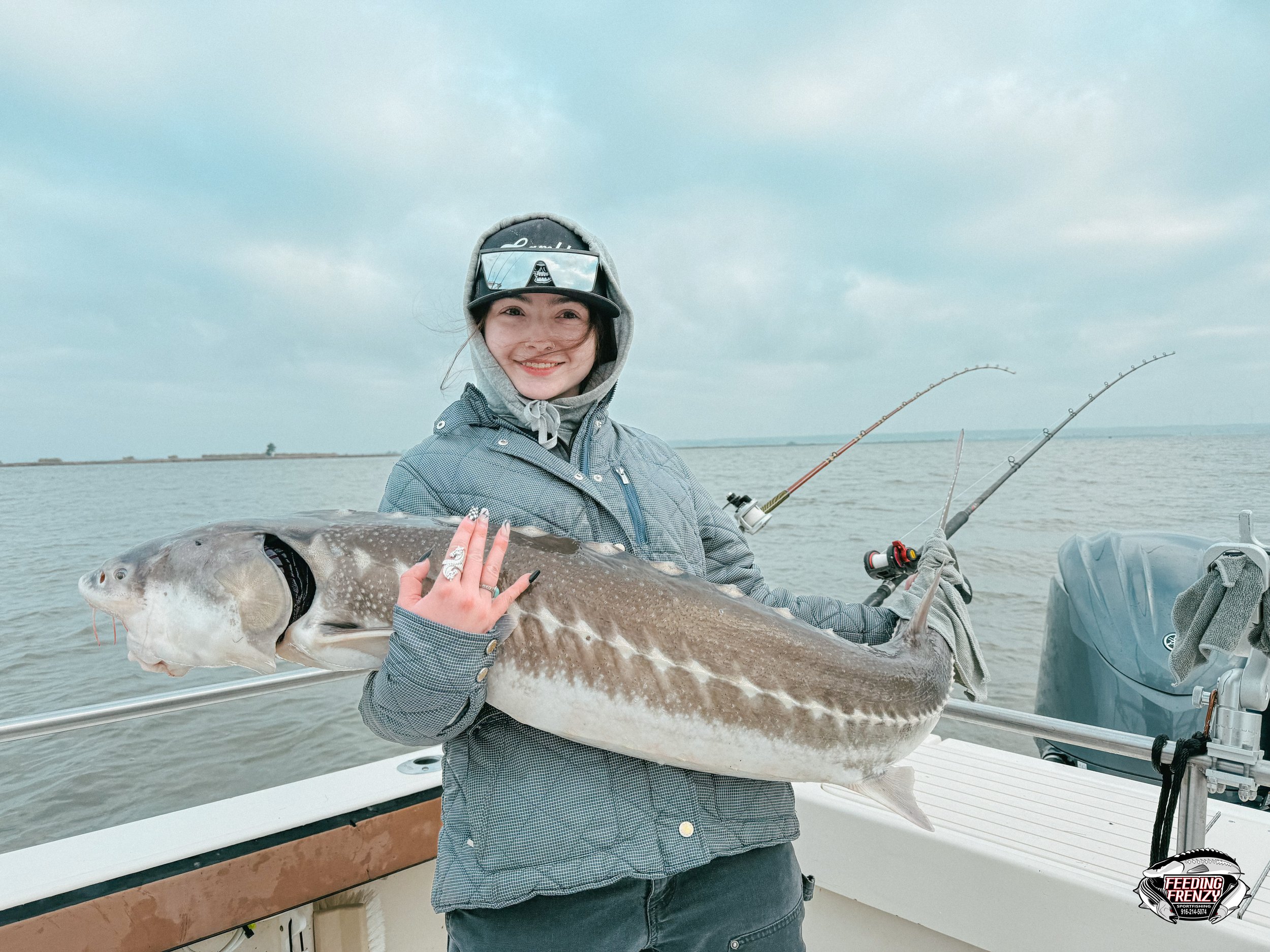 A smiling woman on a boat holding a large fish she caught, with fishing rods in the background on cloudy day.