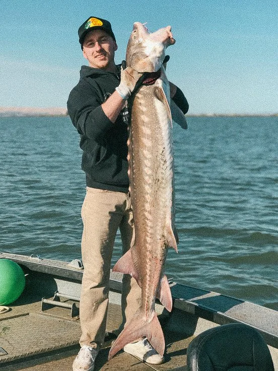 Man wearing a black hoodie, beige pants, and a black cap holding a large fish on a boat in a body of water.