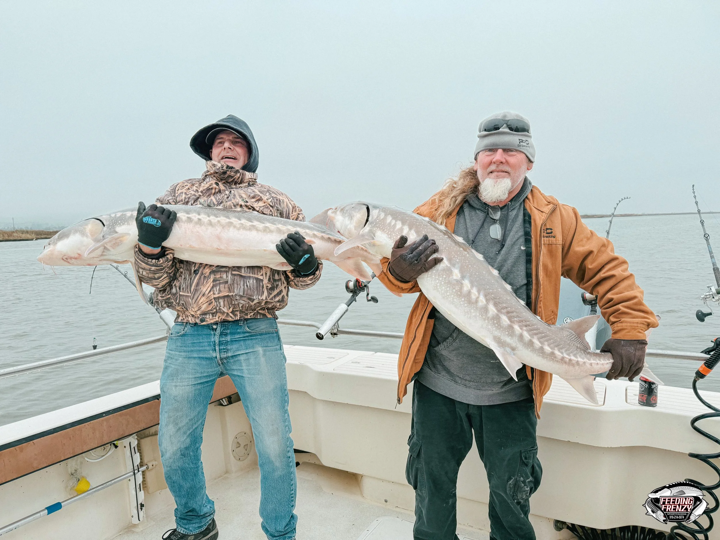 Two men on a boat holding large fish they caught, with fishing rods in the background, on a cloudy day.