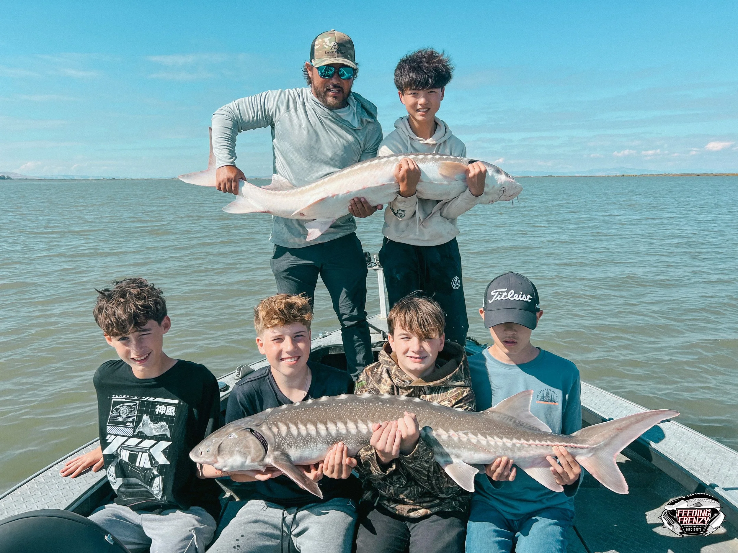A group of six people on a boat holding fish, with two adults standing in the back holding a large fish and four children sitting in front holding smaller fish, on a body of water with clear sky.