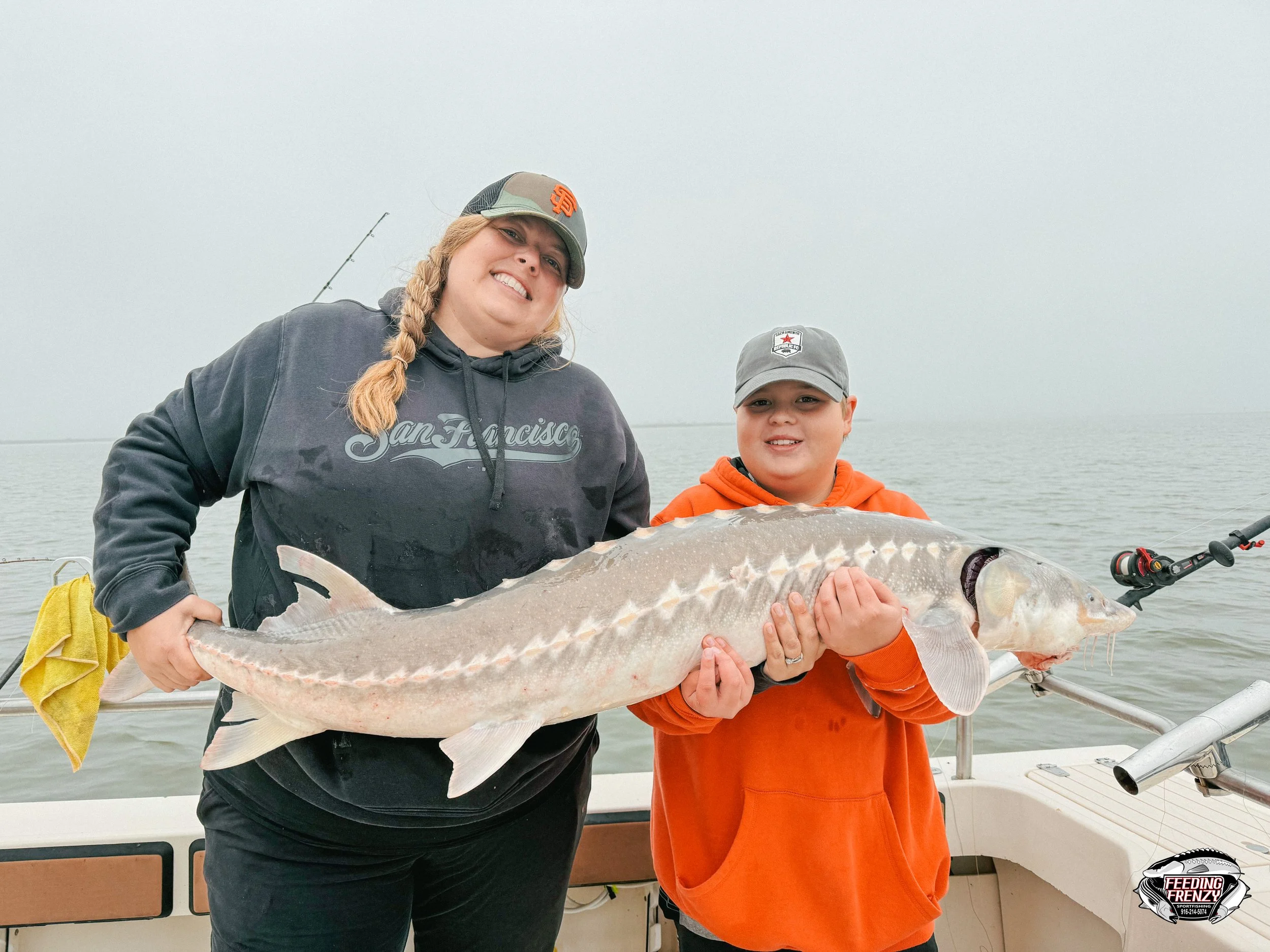 Two people on a boat holding a large fish, with an overcast sky and the ocean in the background.