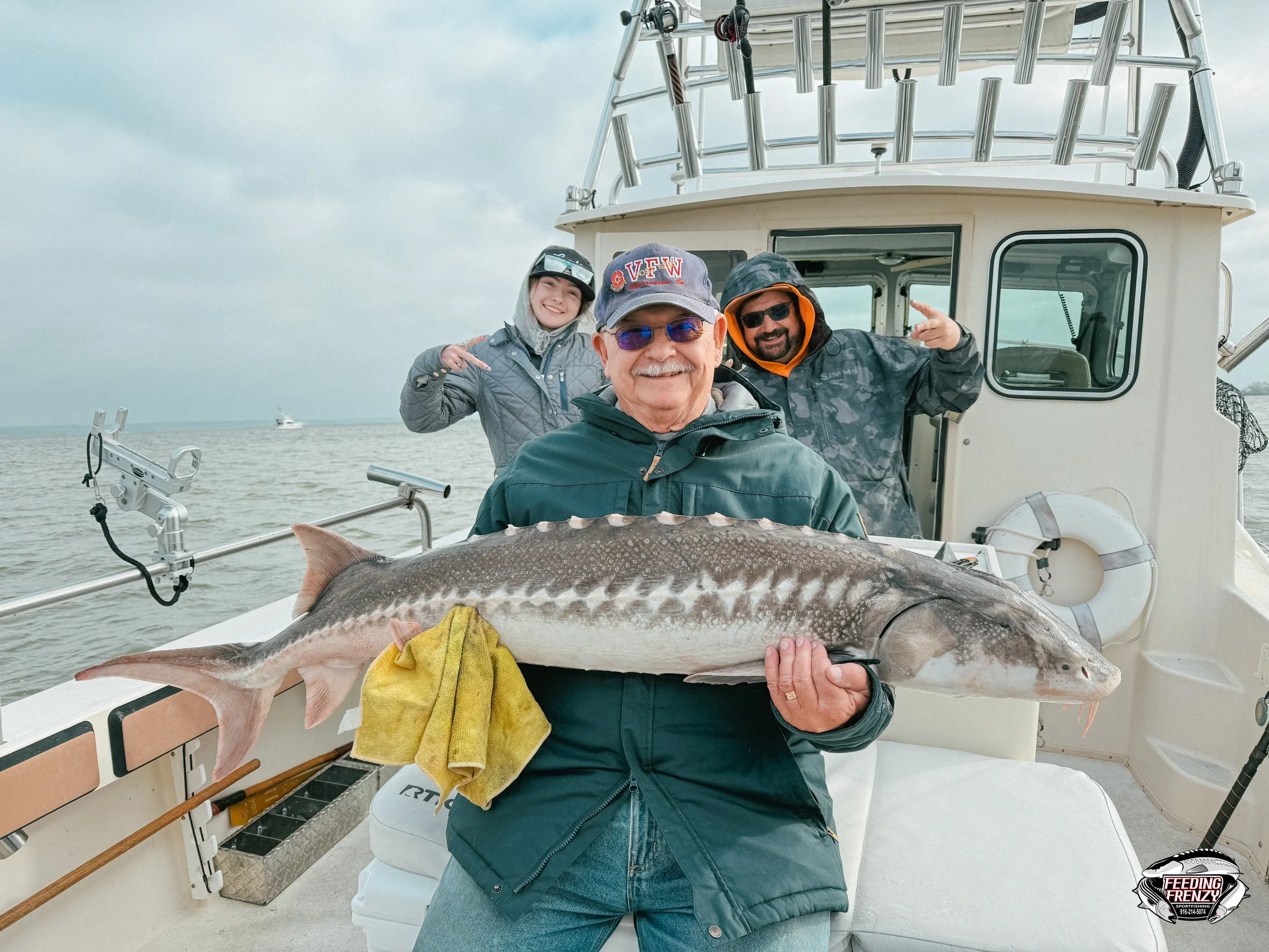 Three men on a boat holding a large fish, with a cloudy sky and other boats in the distance.